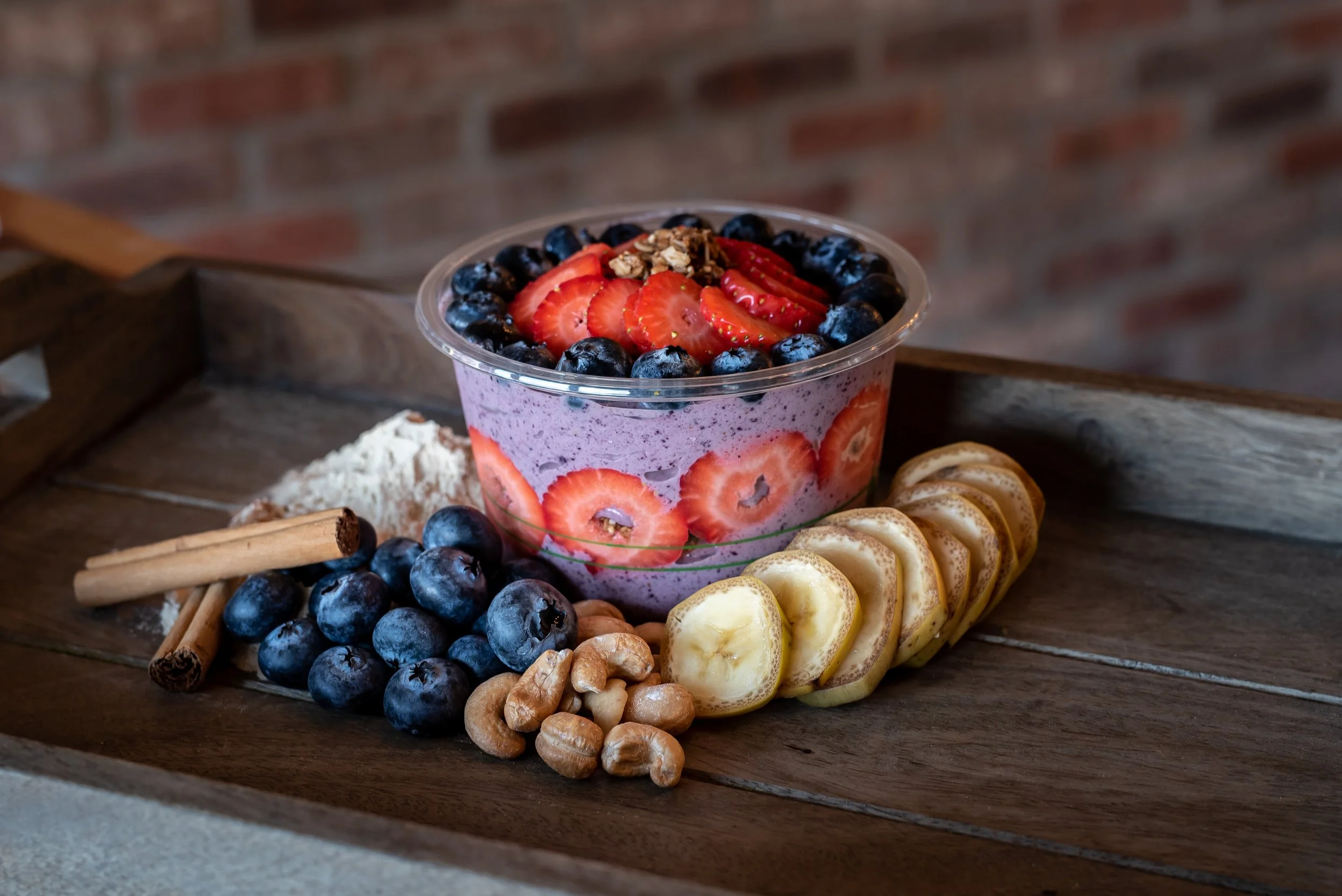 A JuiceKeys bowl filled topped with sliced strawberries and whole blueberries, with a layer of strawberries around the inside of the bowl. Surrounding the bowl are banana slices, blueberries, walnuts, cinnamon sticks, and protein powder on a wooden t