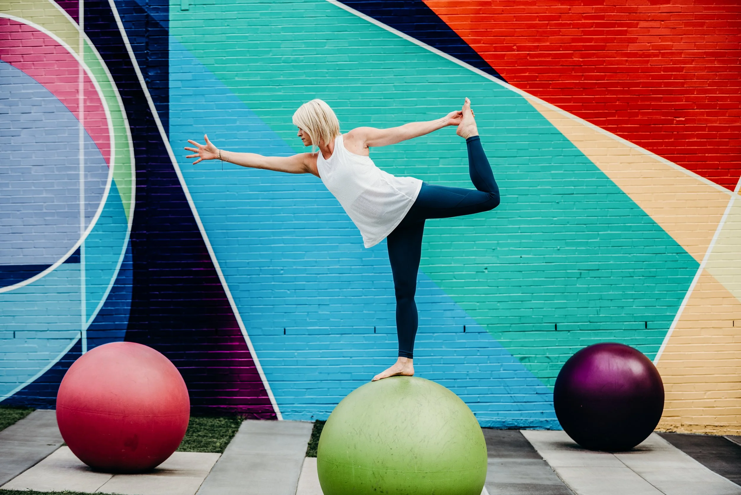 A woman practicing yoga on a green ball in front of a colorful mural, balancing on one foot with her other leg bent behind her, holding her foot with her hand, and extending her arm forward. Photo by Brock Byers 