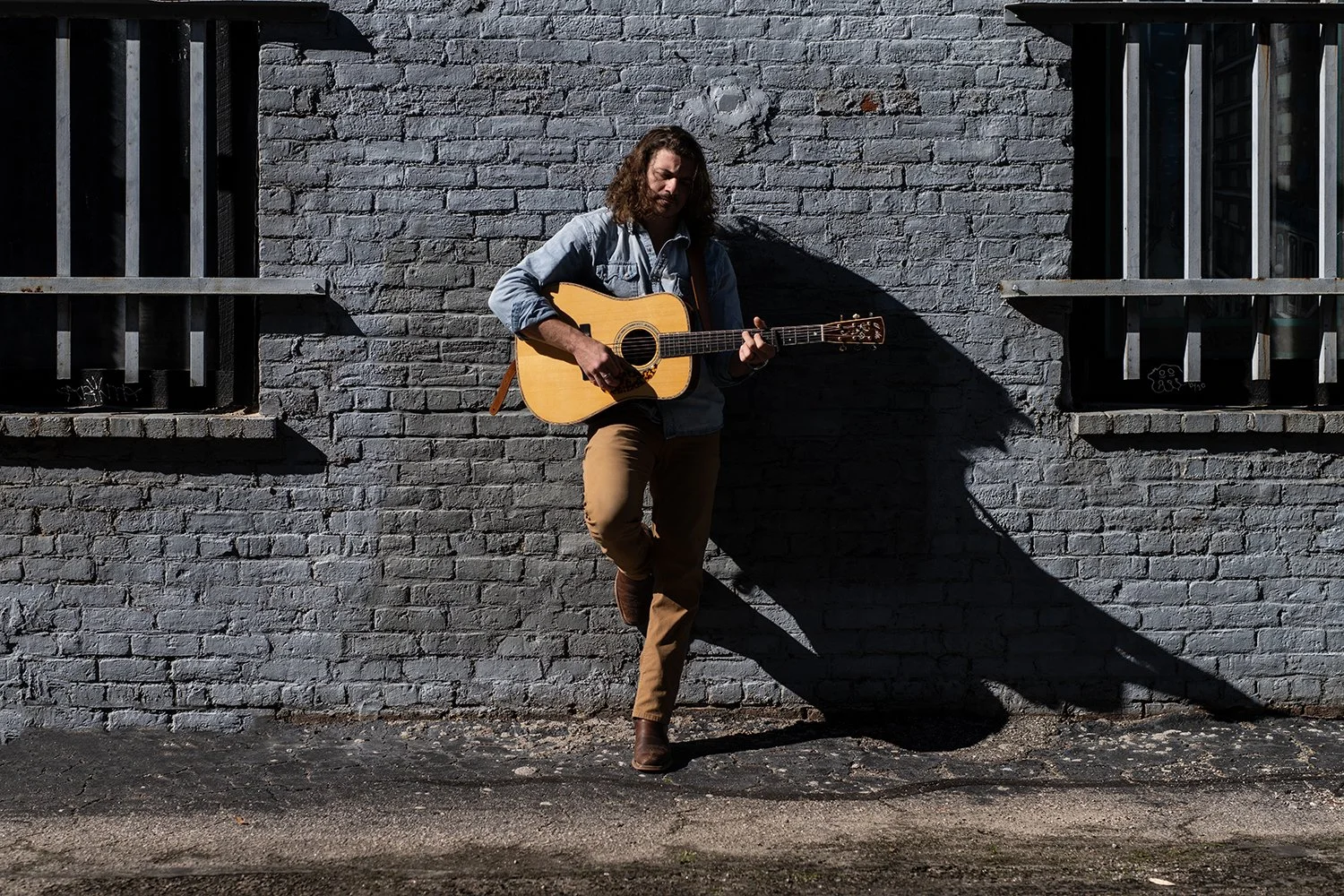 Chris Lewis, musician leaning against a grey brick wall with hard shadows. Photo by Brock Byers 