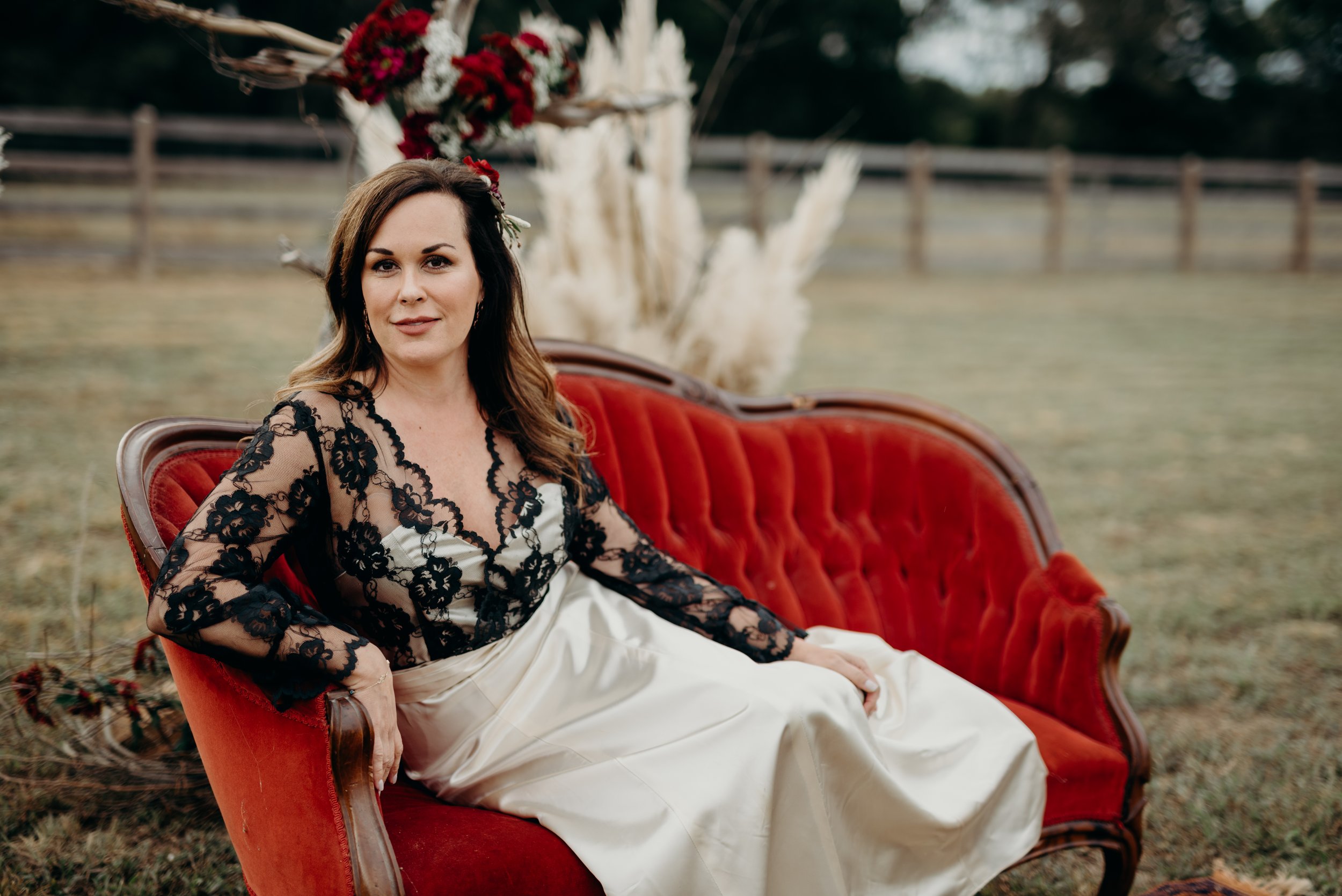A woman sitting on a red vintage sofa outdoors with a decorative floral arrangement behind her, wearing a black lace top and a white skirt, with a calm expression on her face. Photo by Brock Byers 