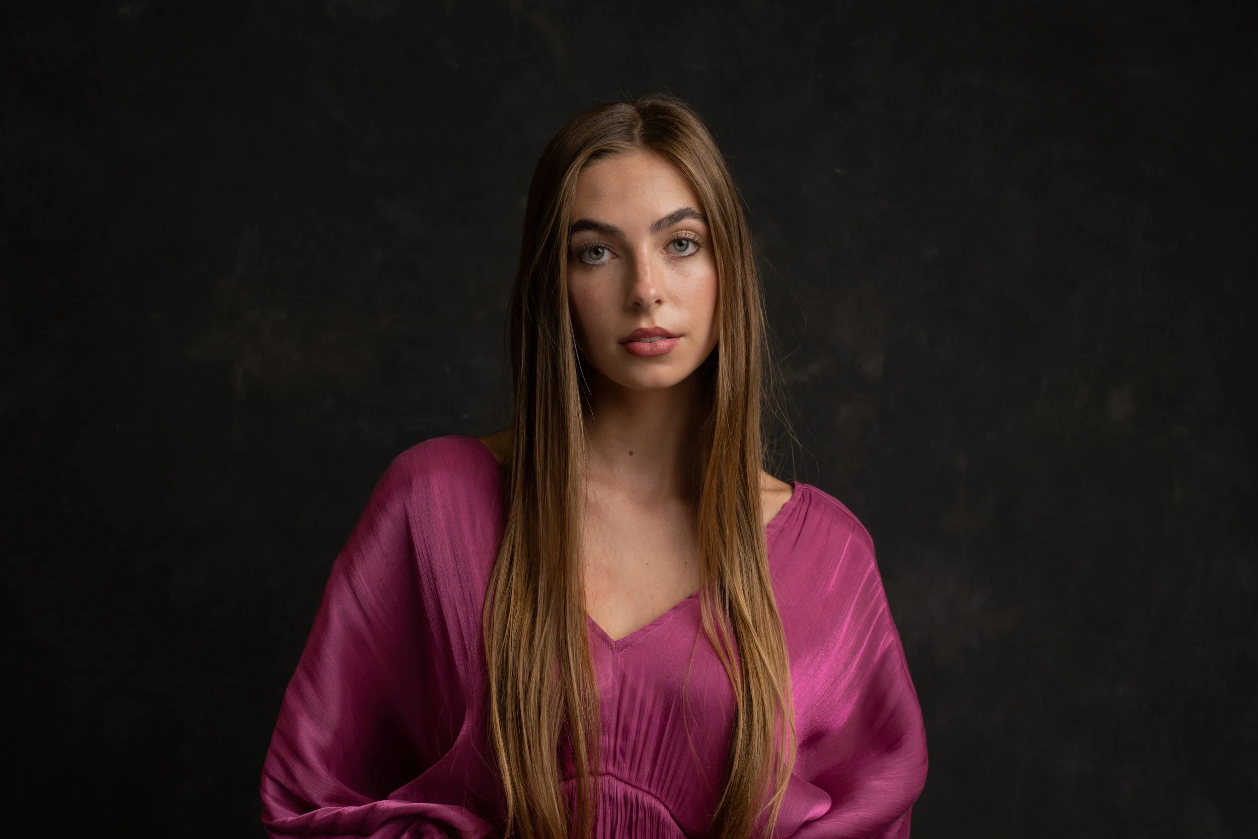 A young woman with long, straight brown hair wearing a purple top, looking directly at the camera against a dark background. Photo by Brock Byers 