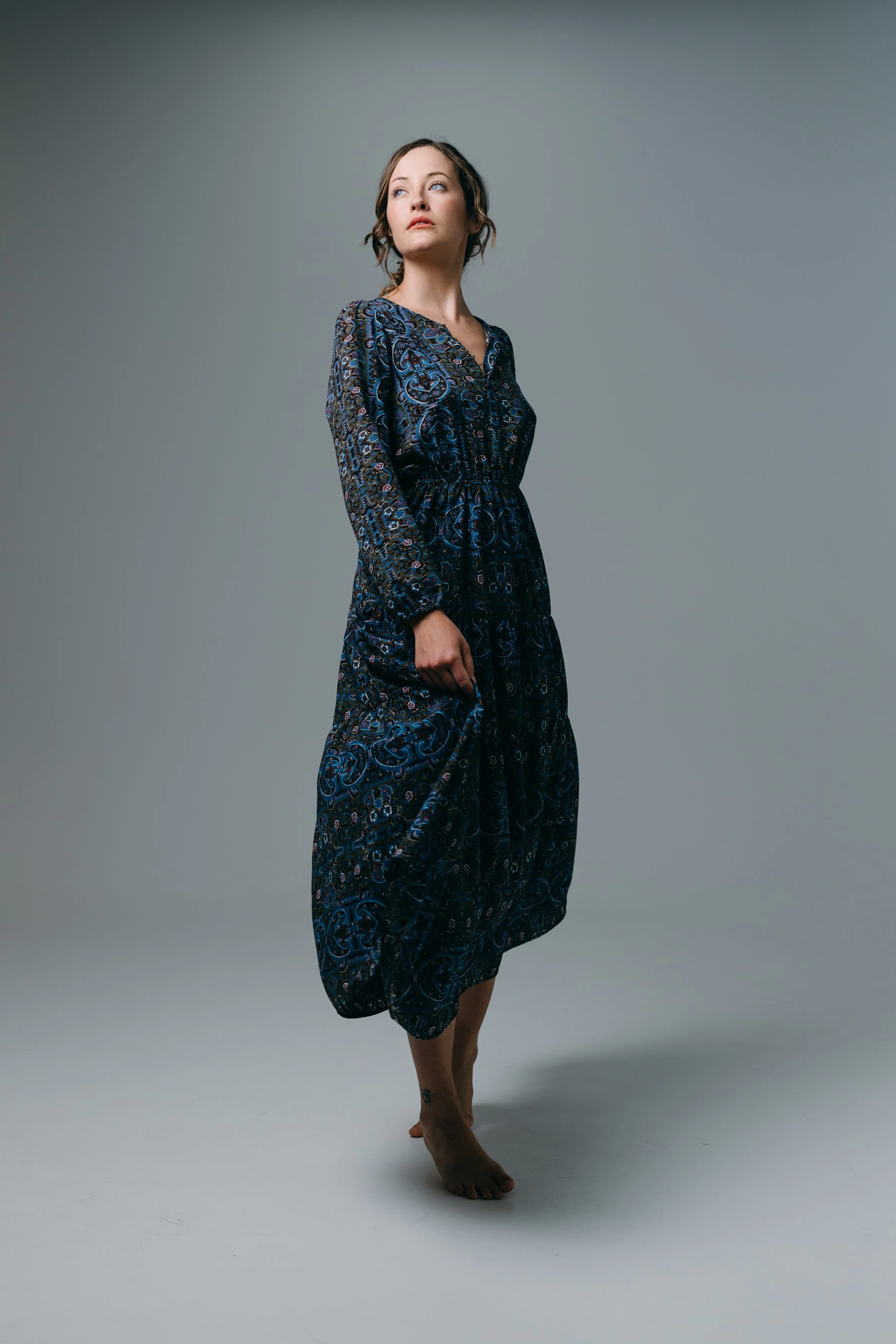 A woman in a dark blue patterned dress standing barefoot on a plain gray background, looking off to the side. Photo by Brock Byers 