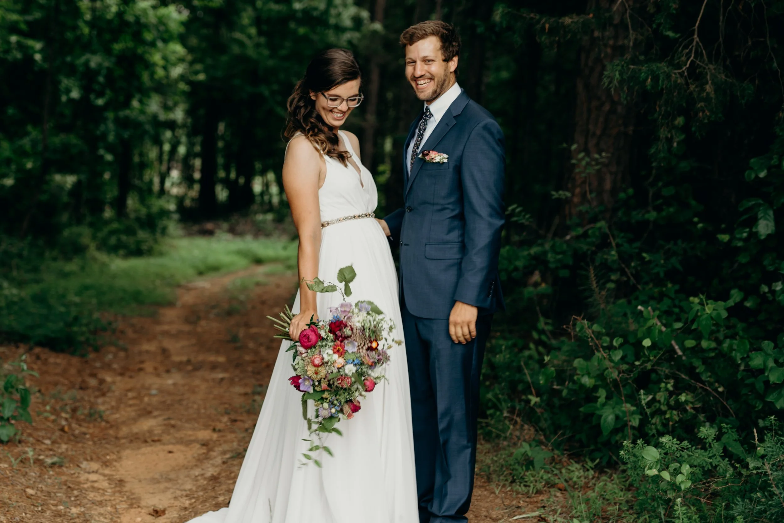 A bride in a white dress holding a colorful bouquet and a groom in a blue suit standing on a forest trail, smiling in a forest setting. Photo by Brock Byers