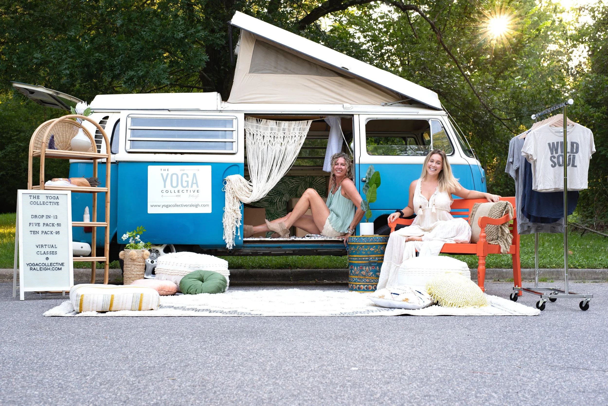 Two women sitting outside a vintage camper van with yoga branding, surrounded by cushions, plants, and clothing on a rack, with a park and trees in the background and sunlight filtering through the trees. Photo by Brock Byers 