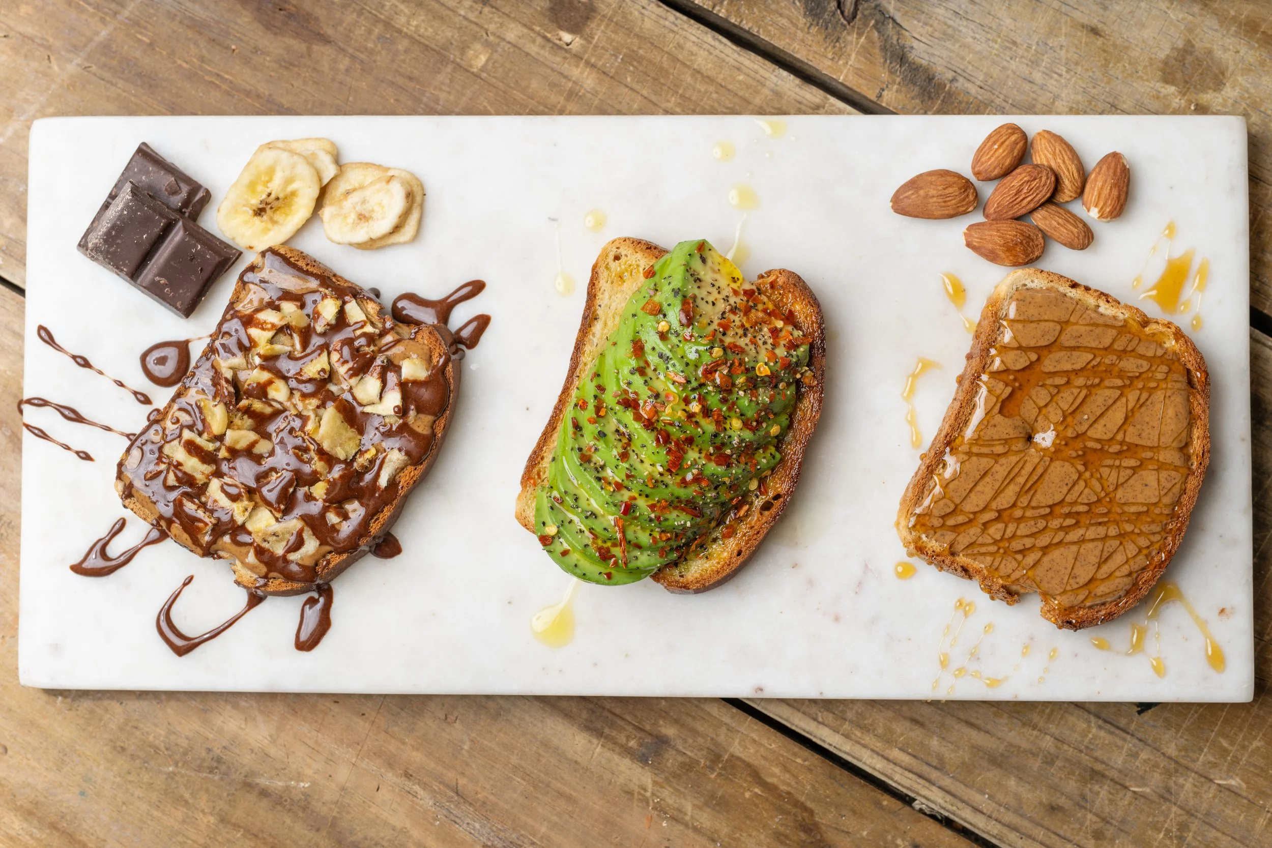 Three slices of toast with different toppings on a white marble serving board, placed on a wooden table. The first slice has banana slices, chocolate pieces, and chocolate drizzle. The second slice has sliced avocado sprinkled with red pepper flakes,