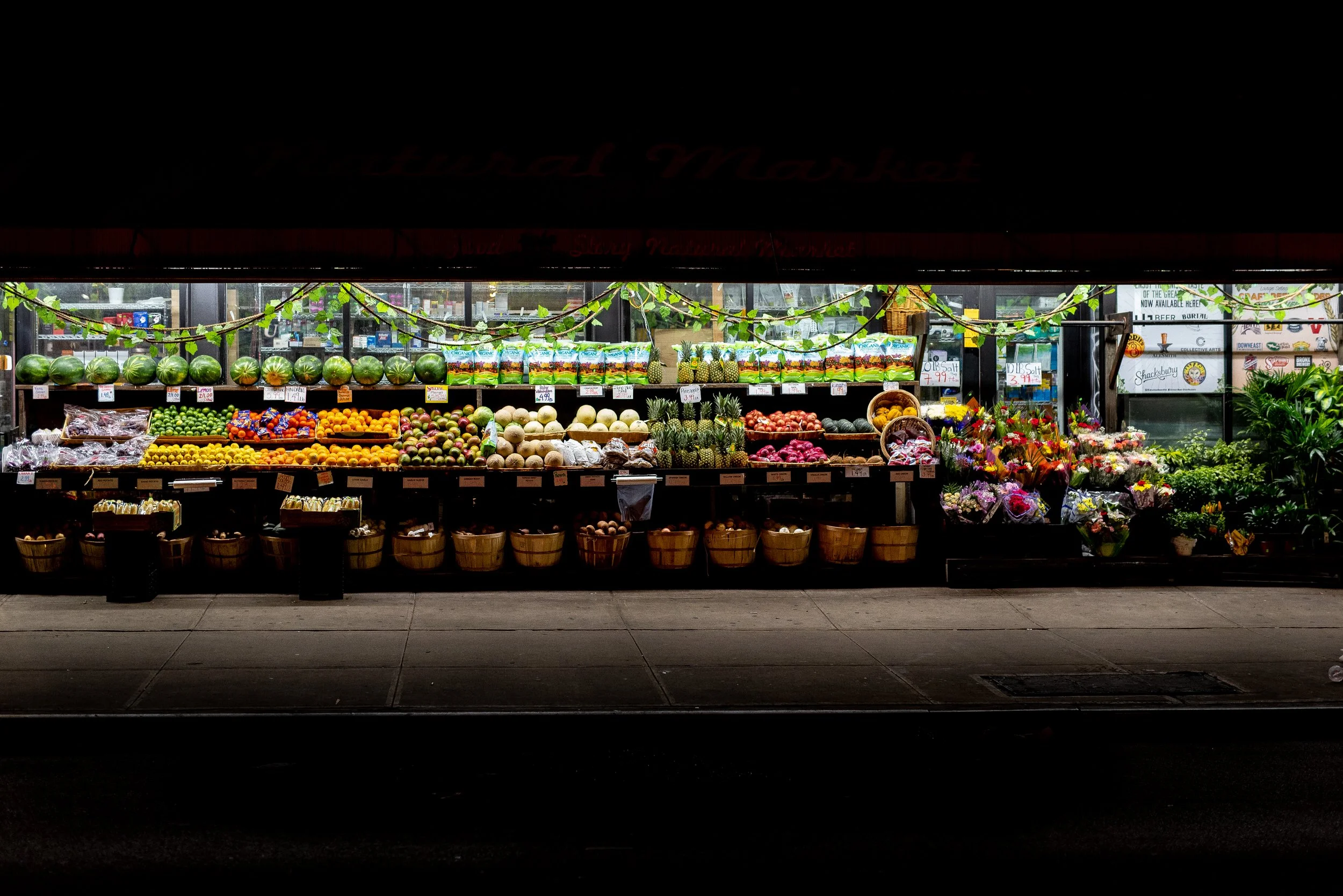 Night view of a Brooklyn, NY supermarket produce section displaying watermelons, pineapples, melons, grapes, apples, and flowers with festive green garlands. Photo by Brock Byers 