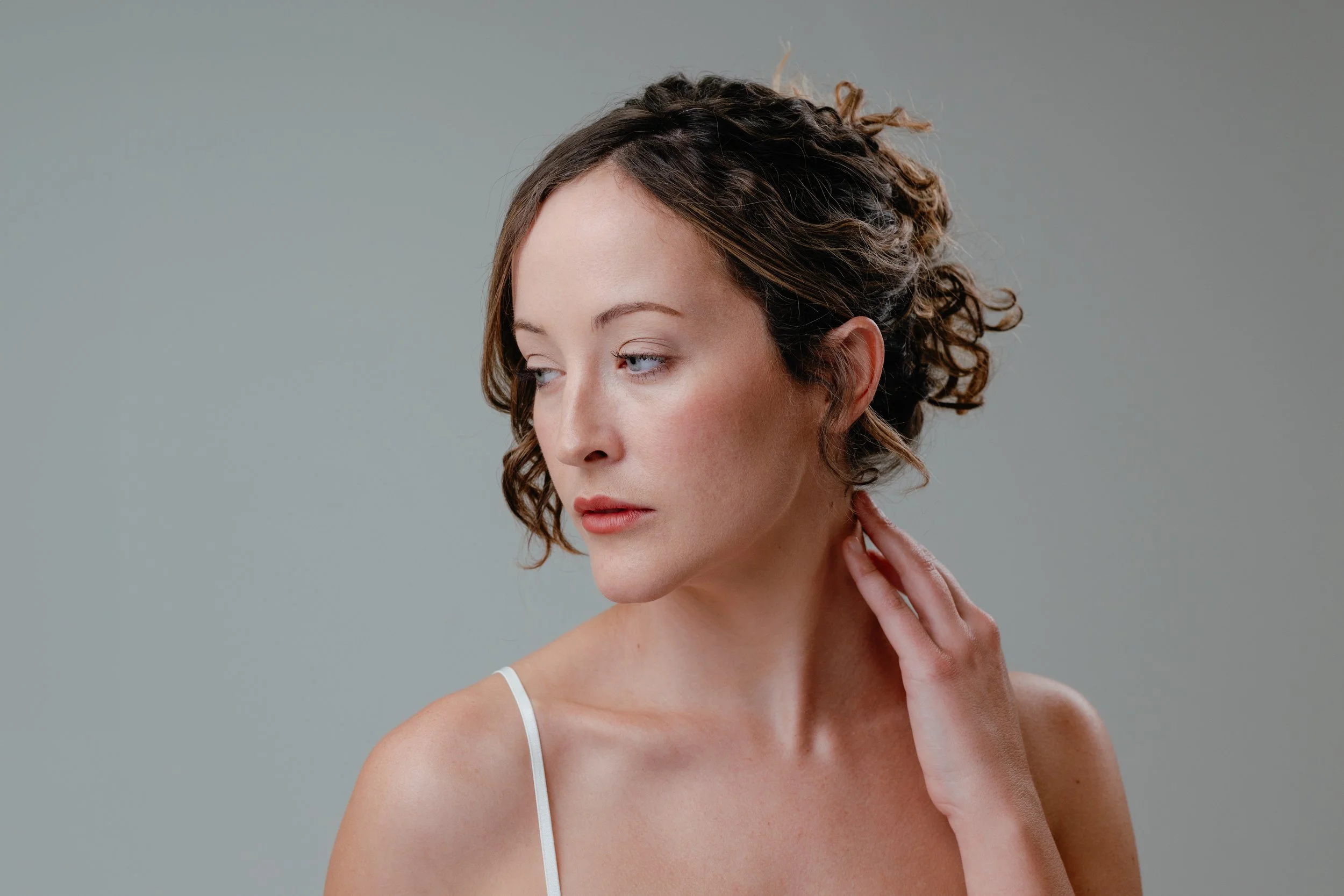 A woman with curly brown hair touching her neck, wearing a white spaghetti strap top, against a plain gray background. Photo by Brock Byers 
