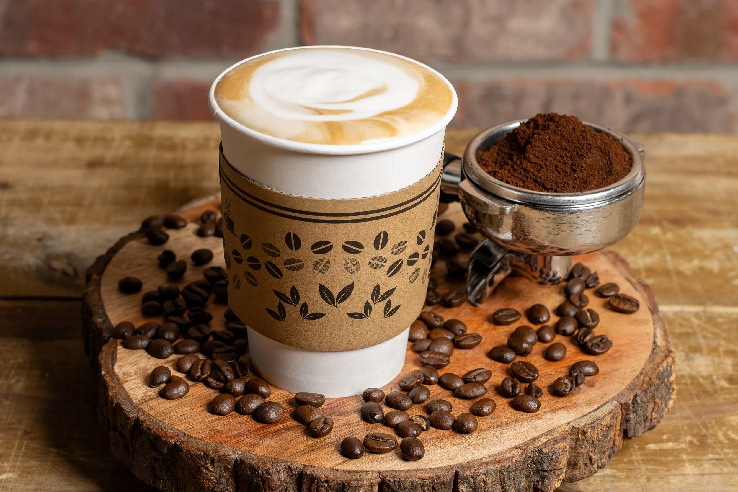 A JuiceKeys to-go coffee cup with latte art, placed on a wooden surface covered with coffee beans, next to a small metal container filled with ground coffee on a round wooden serving board. Photo by Brock Byers 