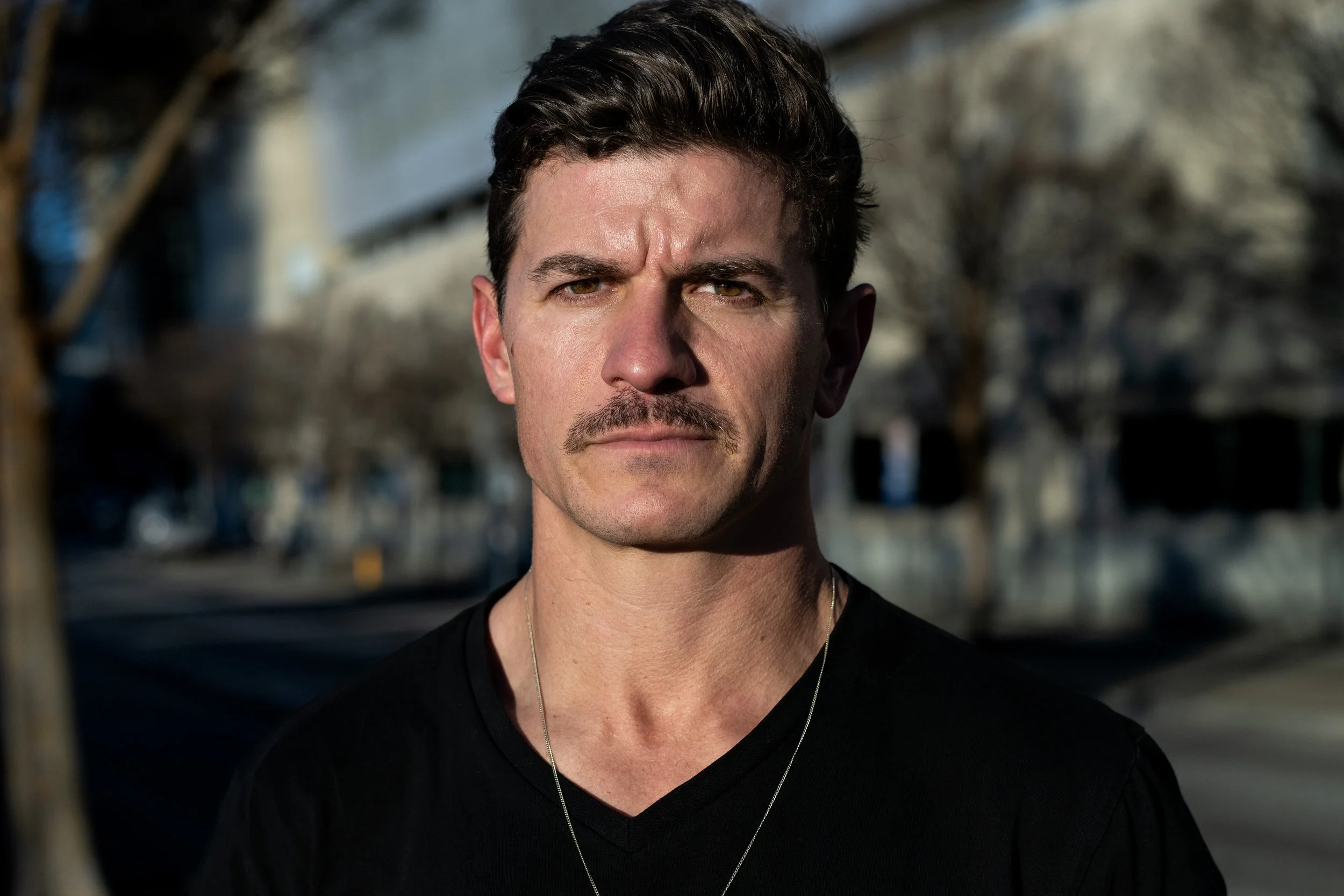 Close-up of a man with dark hair and a mustache, wearing a black shirt and a silver necklace, outdoors in a sunny urban setting with blurred trees and buildings in the background. Photo by Brock Byers 