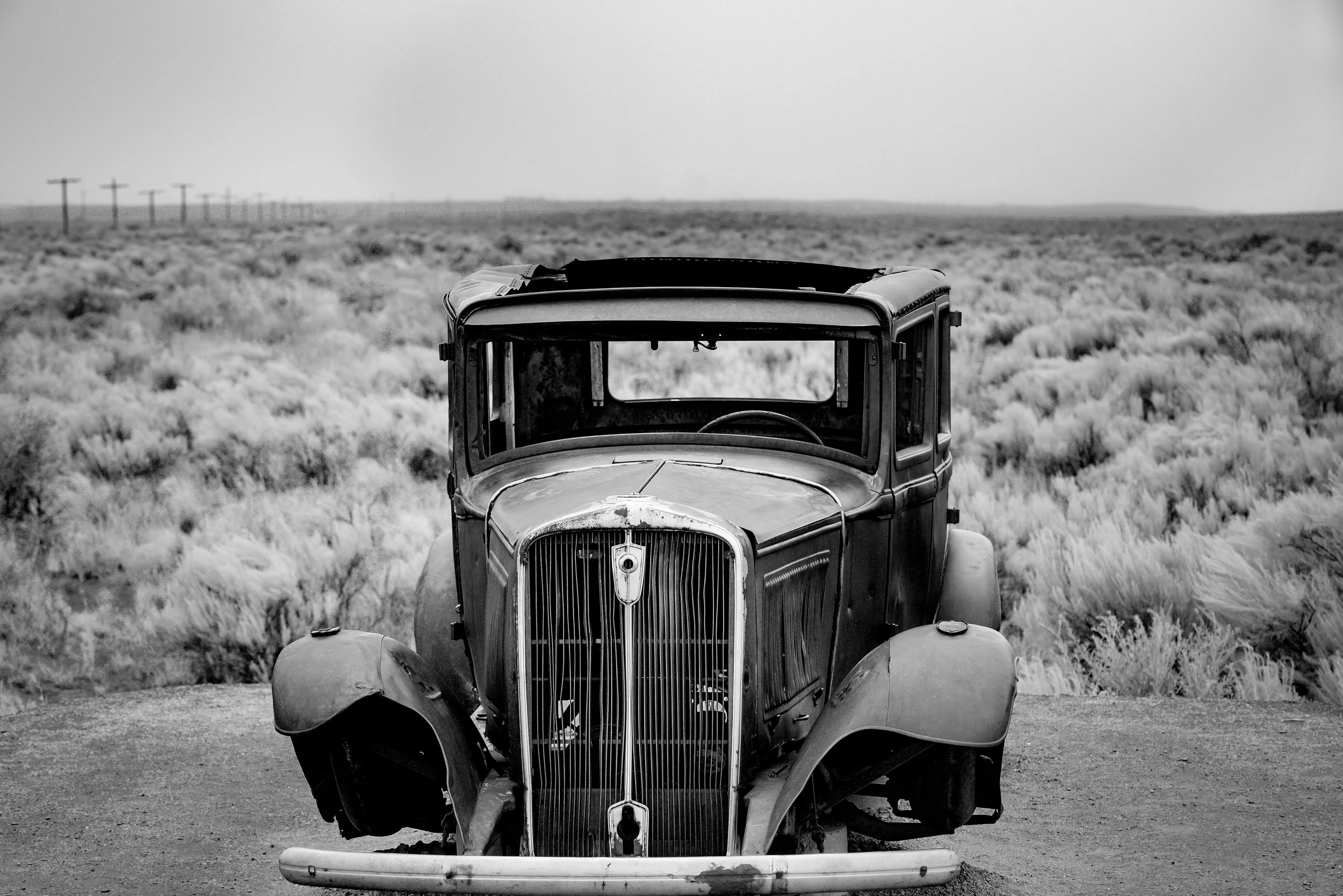 Black and white photograph of an old, abandoned vintage car in a desert landscape with sparse vegetation and power lines in the distance. Photo by Brock Byers 