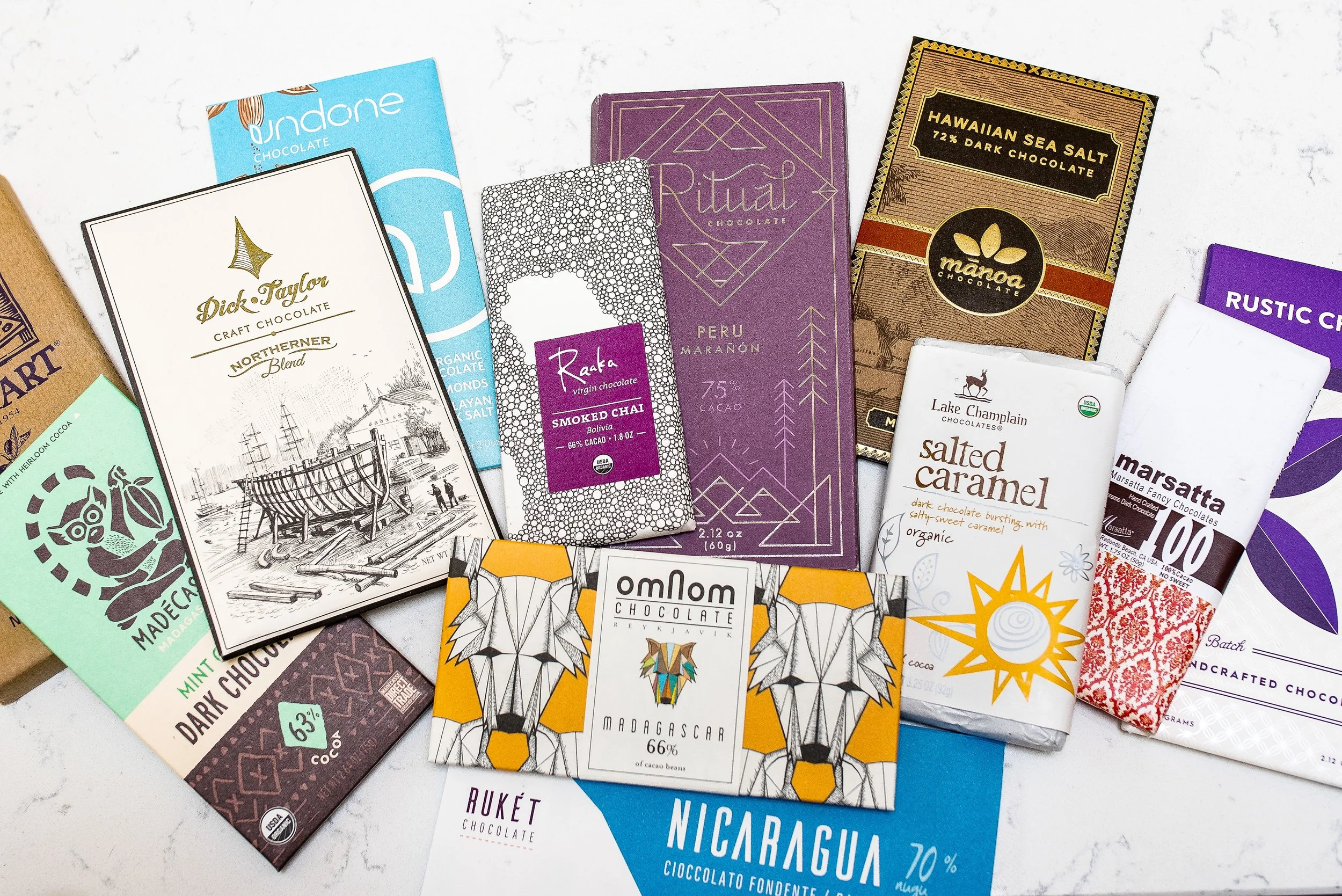 An assortment of chocolate bars with various brand labels, flavors, and packaging designs scattered on a light surface. Photo by Brock Byers 