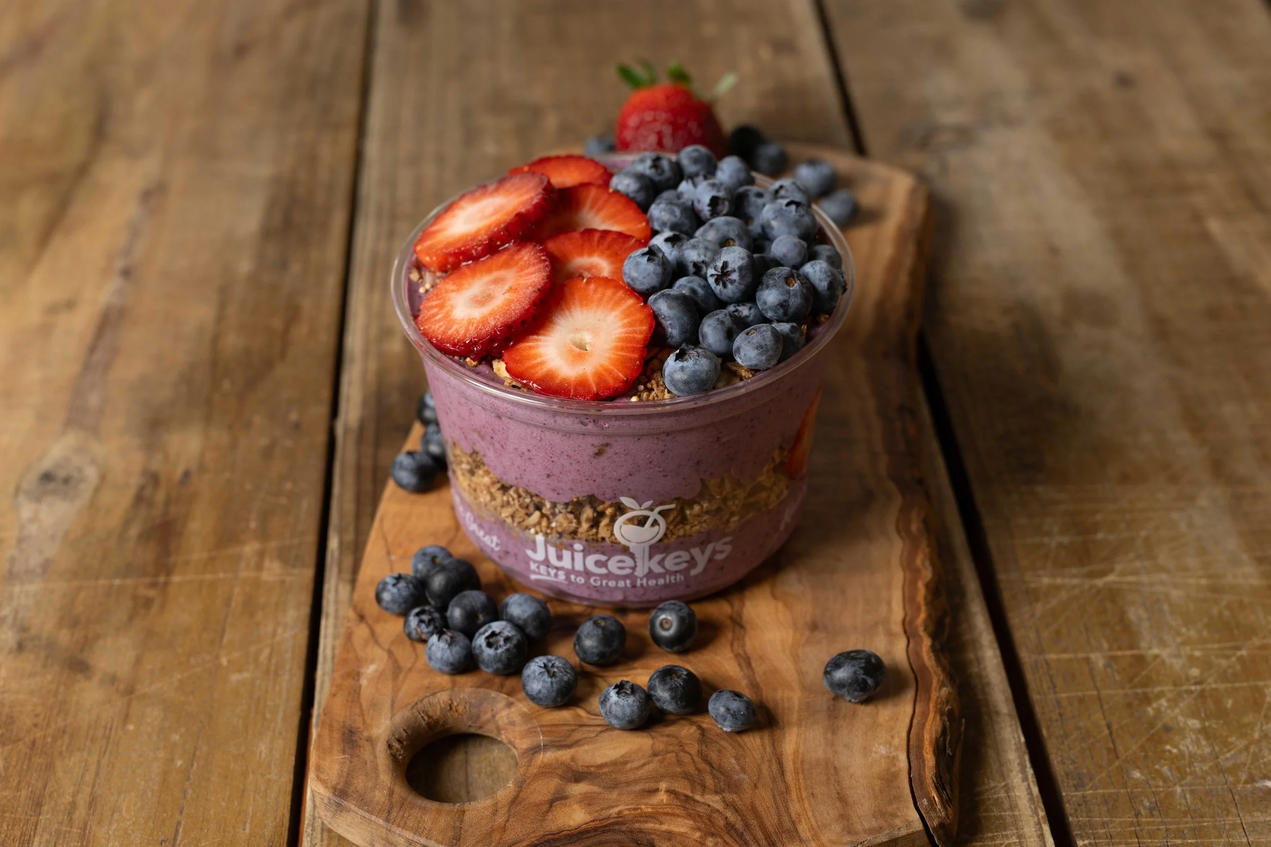 A layered JuiceKeys bowl with sliced strawberries and blueberries on top, served in a plastic bowl on a wooden cutting board with scattered blueberries around. Photo by Brock Byers 