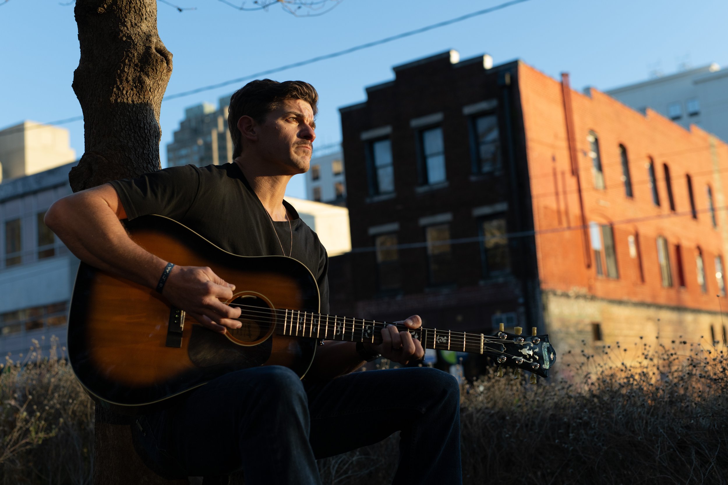 A man sitting outdoors playing an acoustic guitar, with buildings and a clear sky in the background during late afternoon or early evening. Photo by Brock Byers 