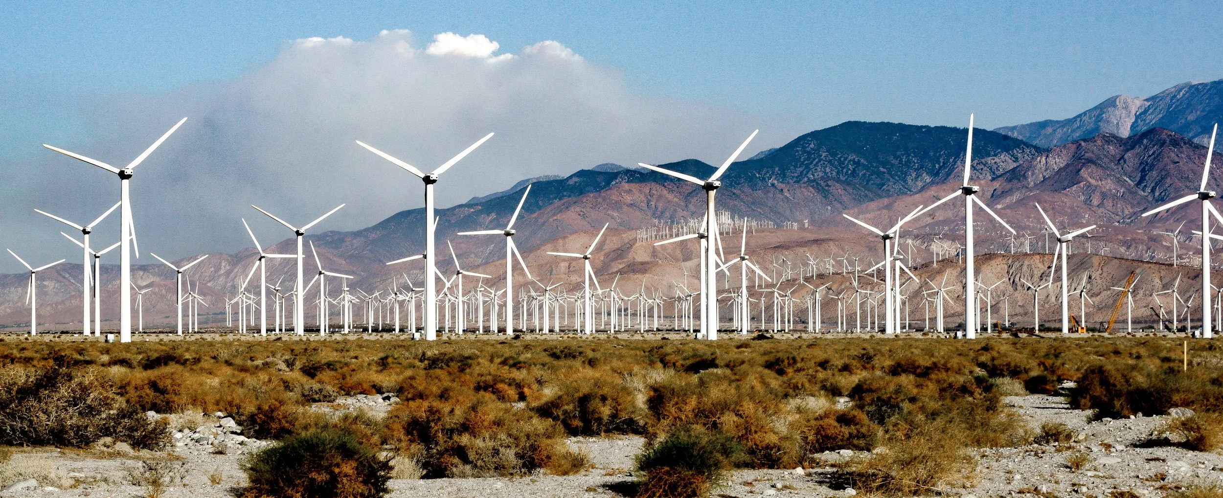 A desert landscape with numerous white wind turbines against a backdrop of mountains and a partly cloudy sky. Photo by Brock Byers