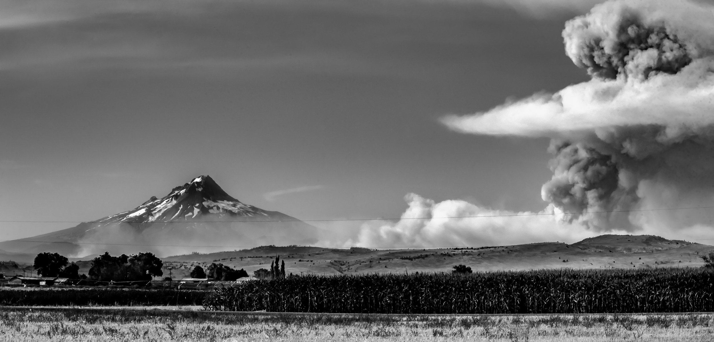 A black and white photo of Mount Hood on the left and a wildfire with large ash clouds on the right, over farmland and hills in the foreground. Photo by Brock Byers 