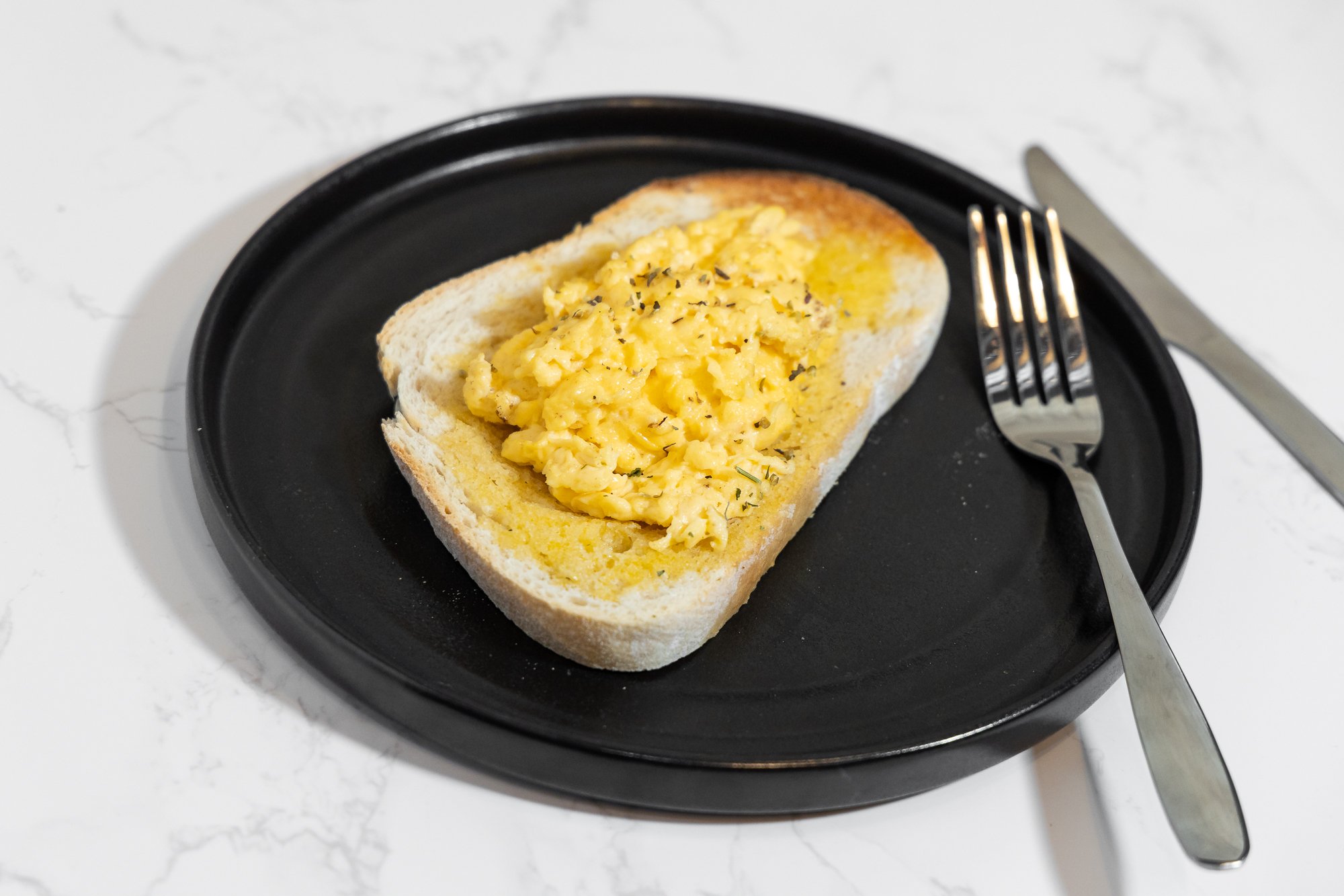 Scrambled eggs on a toasted bread slice on a black plate with a fork and knife on a white marble surface.