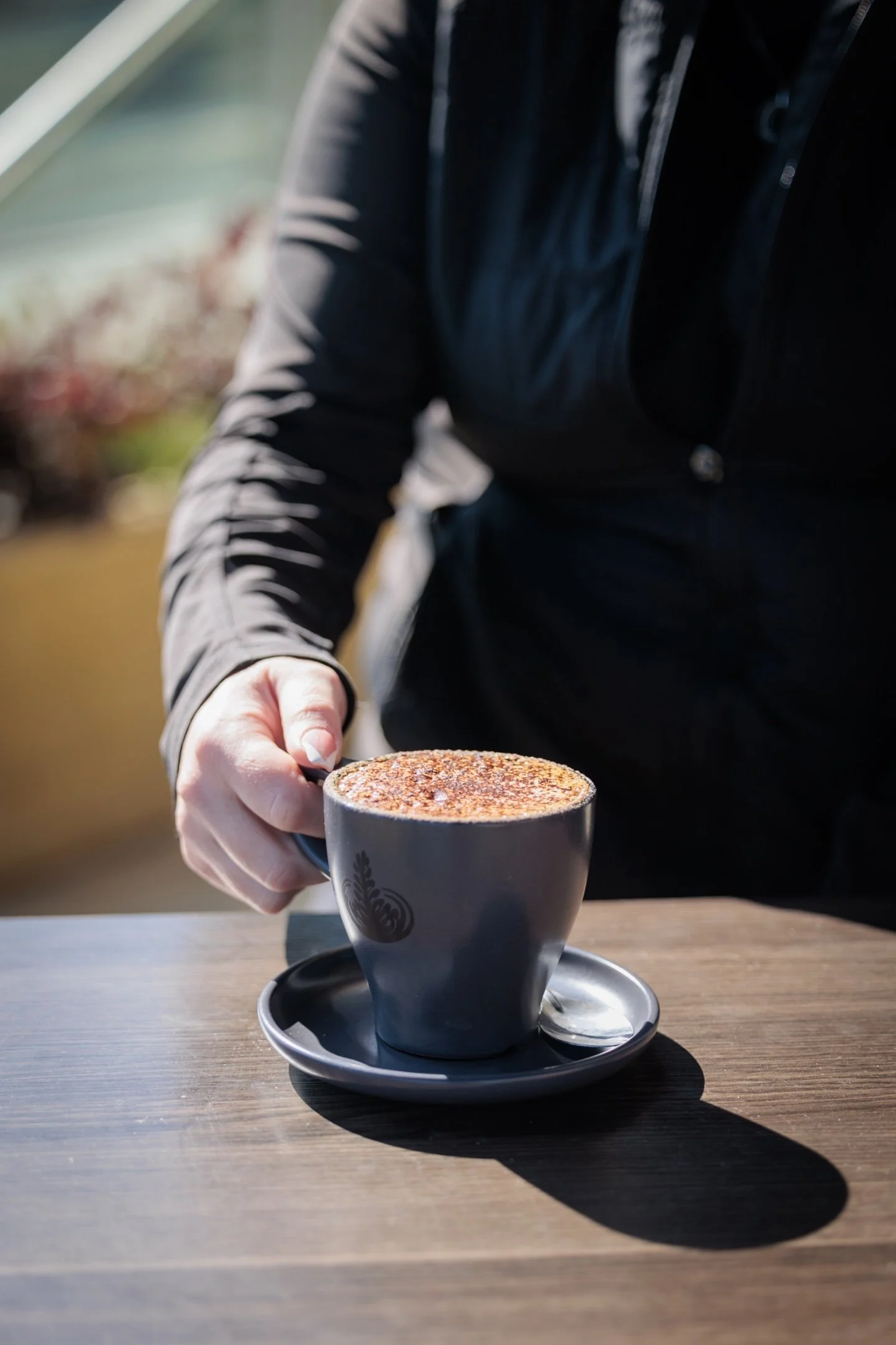 Person holding a black coffee cup with frothy topping, sitting at a wooden table outdoors.