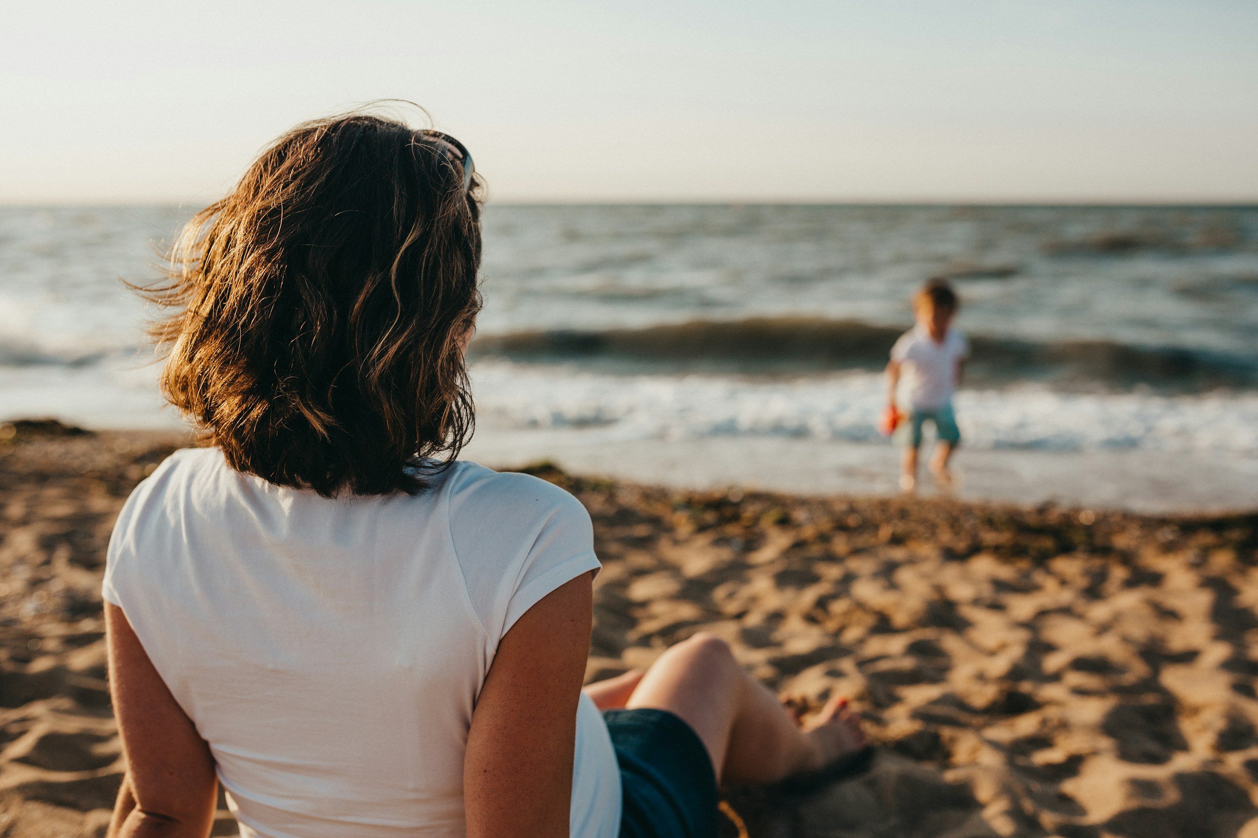 A mother sitting on the sand looking toward her young child playing in the ocean waves, capturing the emotional dissonance and overwhelm of summer parenting.