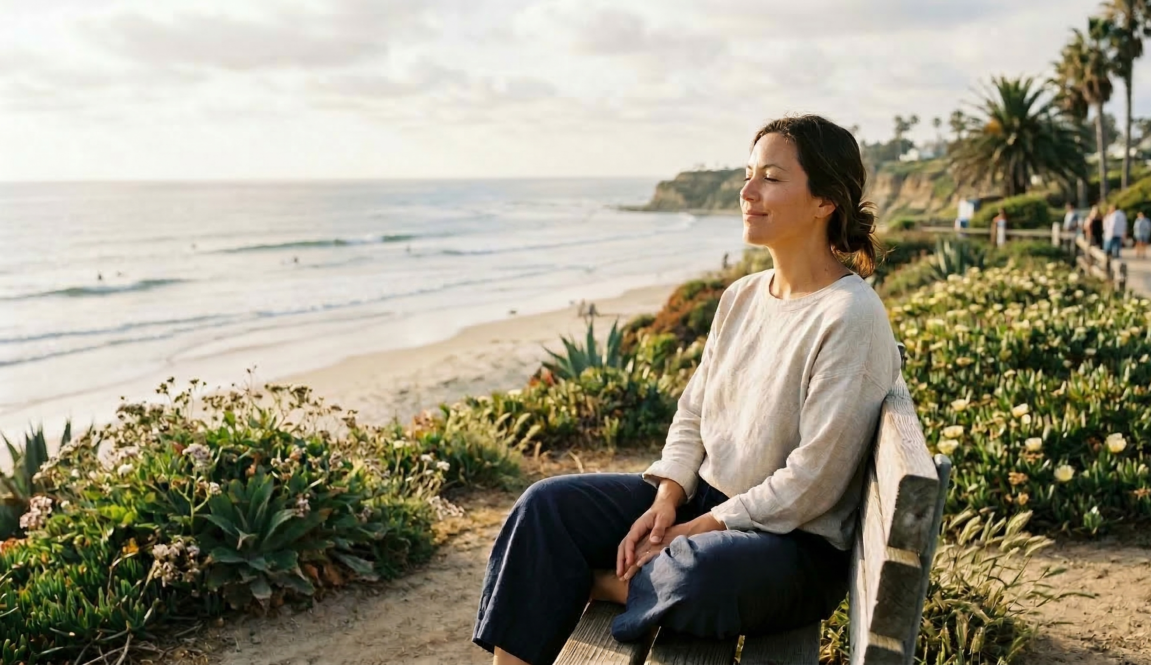 A woman sitting on a bench overlooking the ocean in Encinitas, taking a deep breath and experiencing a moment of calm, representing postpartum support and maternal therapy in North County San Diego.