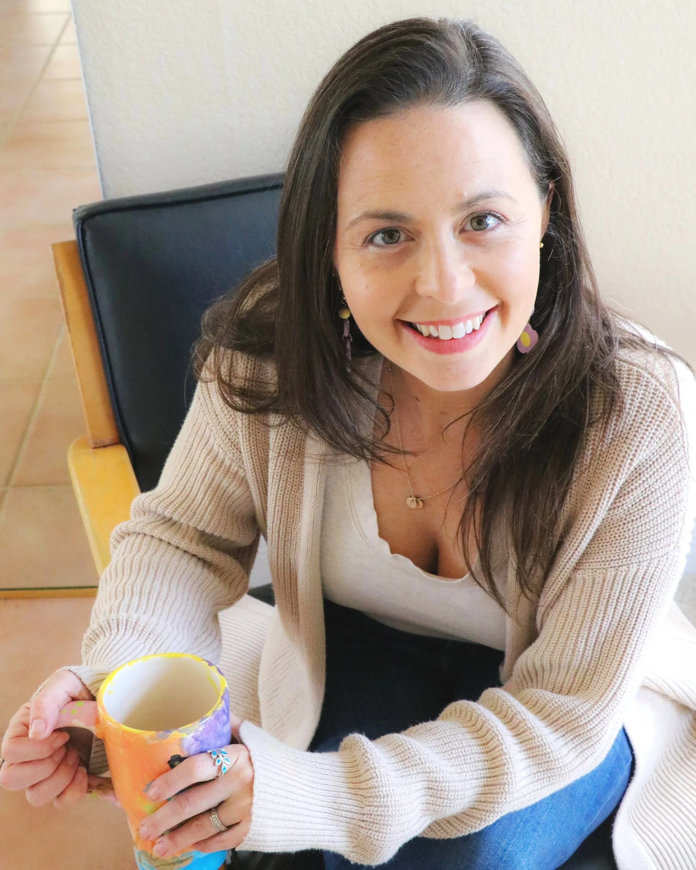 Ellie Messinger-Adams with brown hair and blue eyes smiling, wearing a beige cardigan and white top, sitting on a black chair, holding a colorful mug.