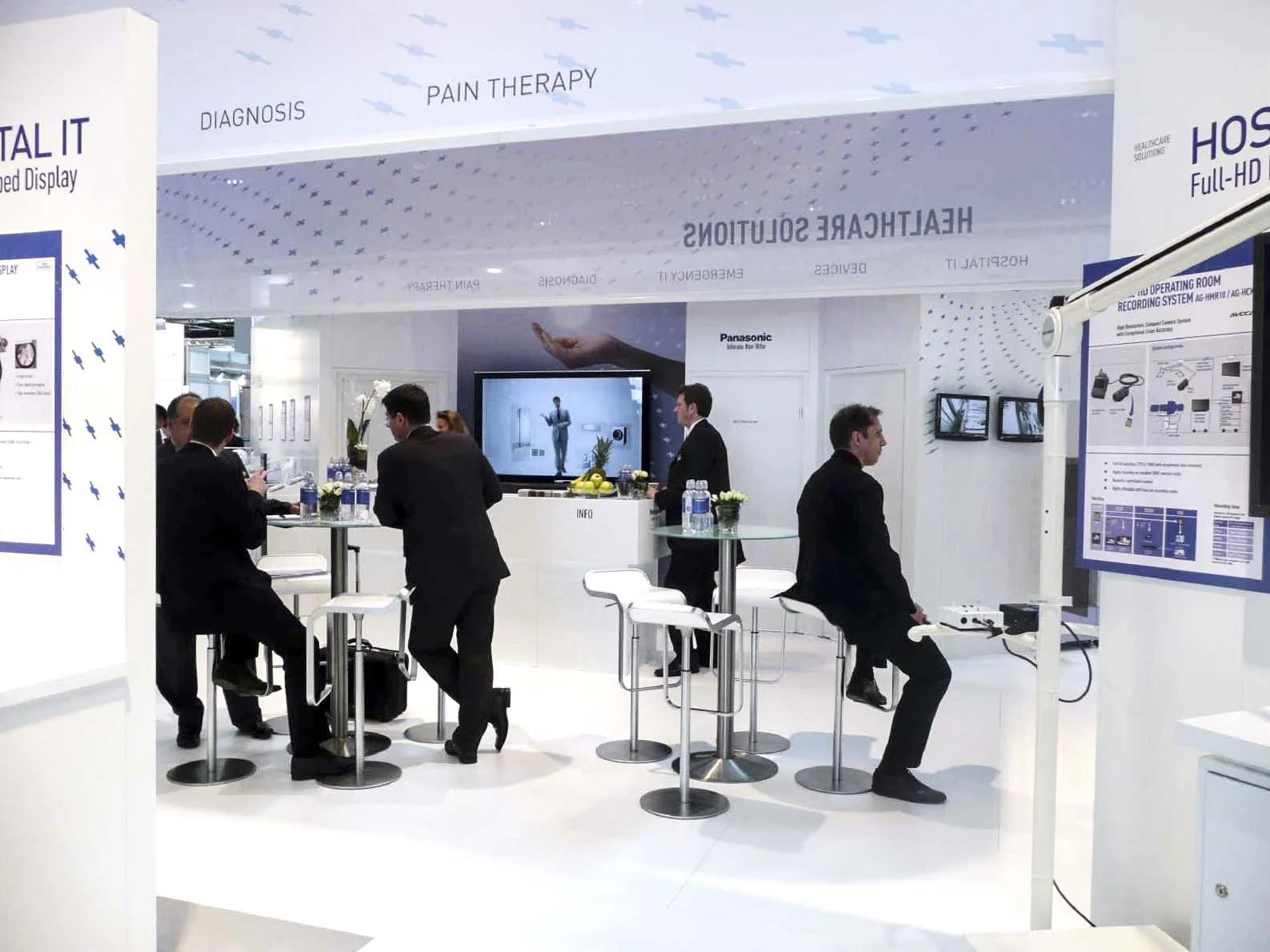 Innenansicht des Panasonic Healthcare-Messestands mit weißer Gestaltung, Themenbereichen wie Diagnosis und Pain Therapy und Besuchern im Gespräch
