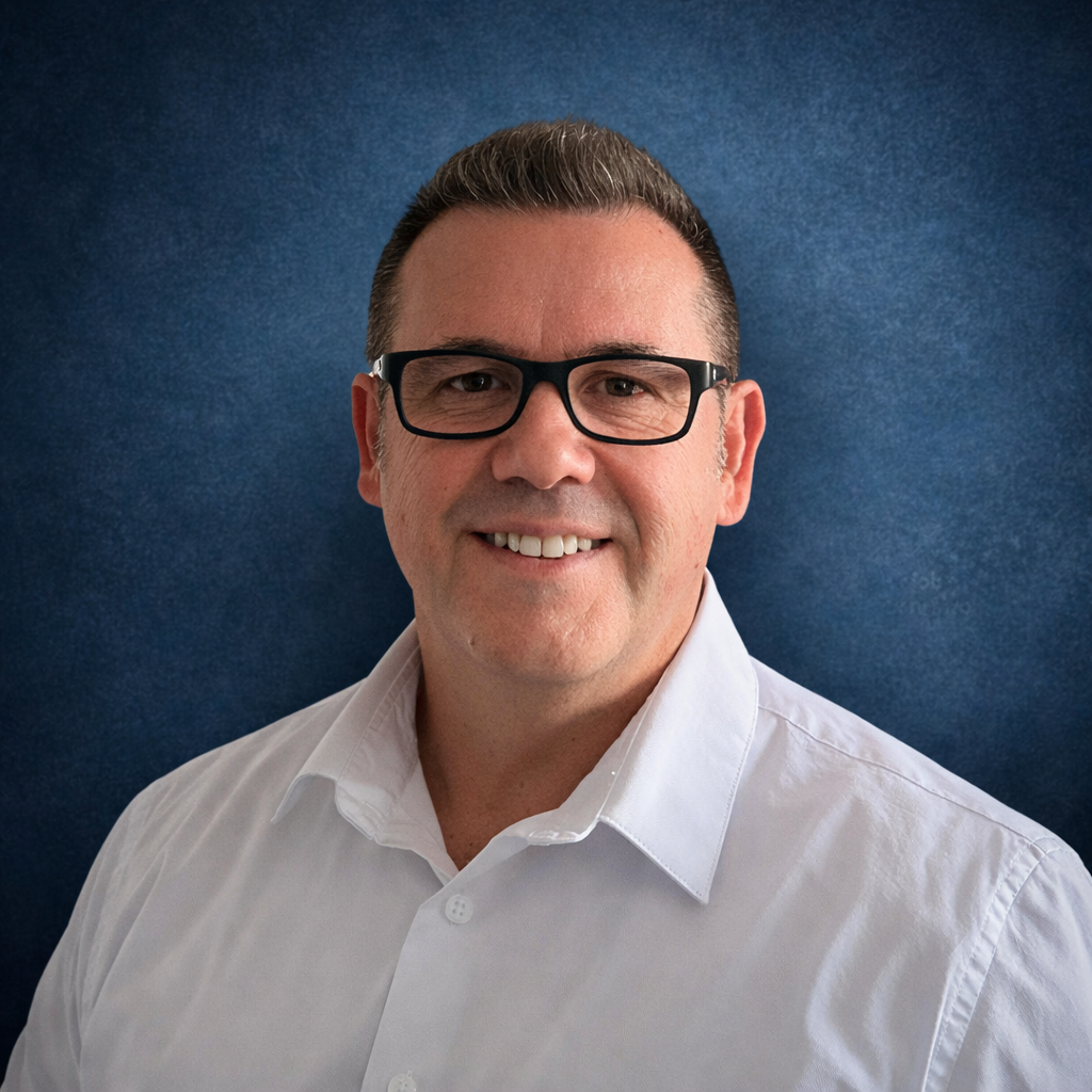 Portrait d'un homme souriant portant des lunettes noires et une chemise blanche, avec un fond bleu