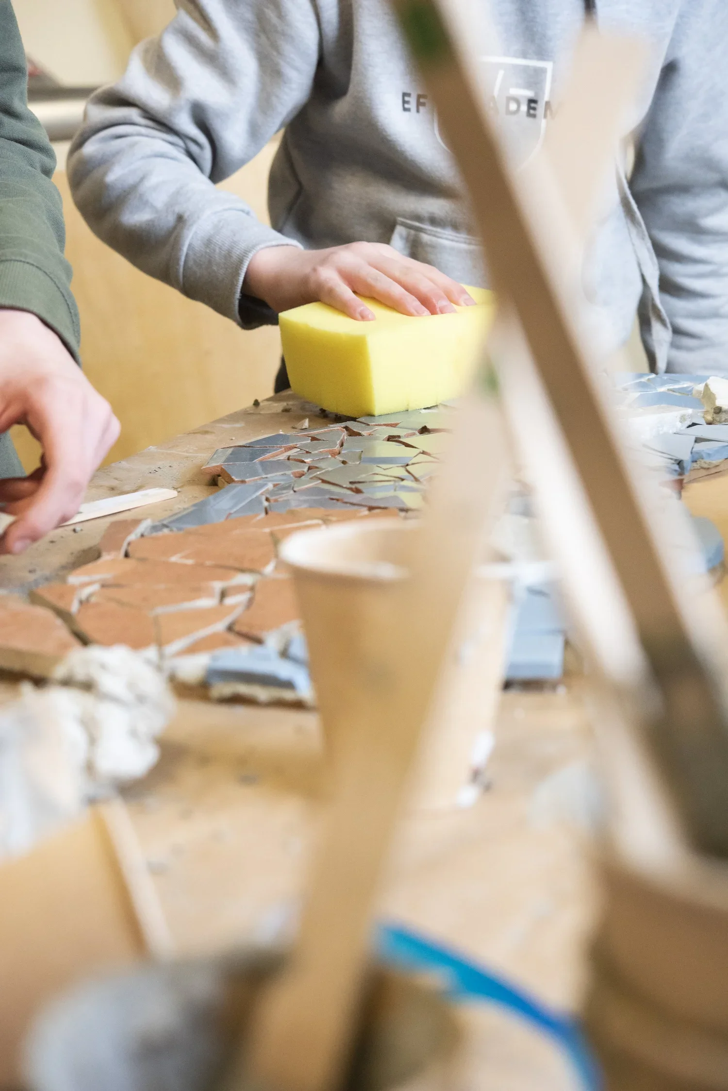 People working on a mosaic art project at a table with broken ceramic tiles and tools, with a person pressing yellow sponge onto the surface.