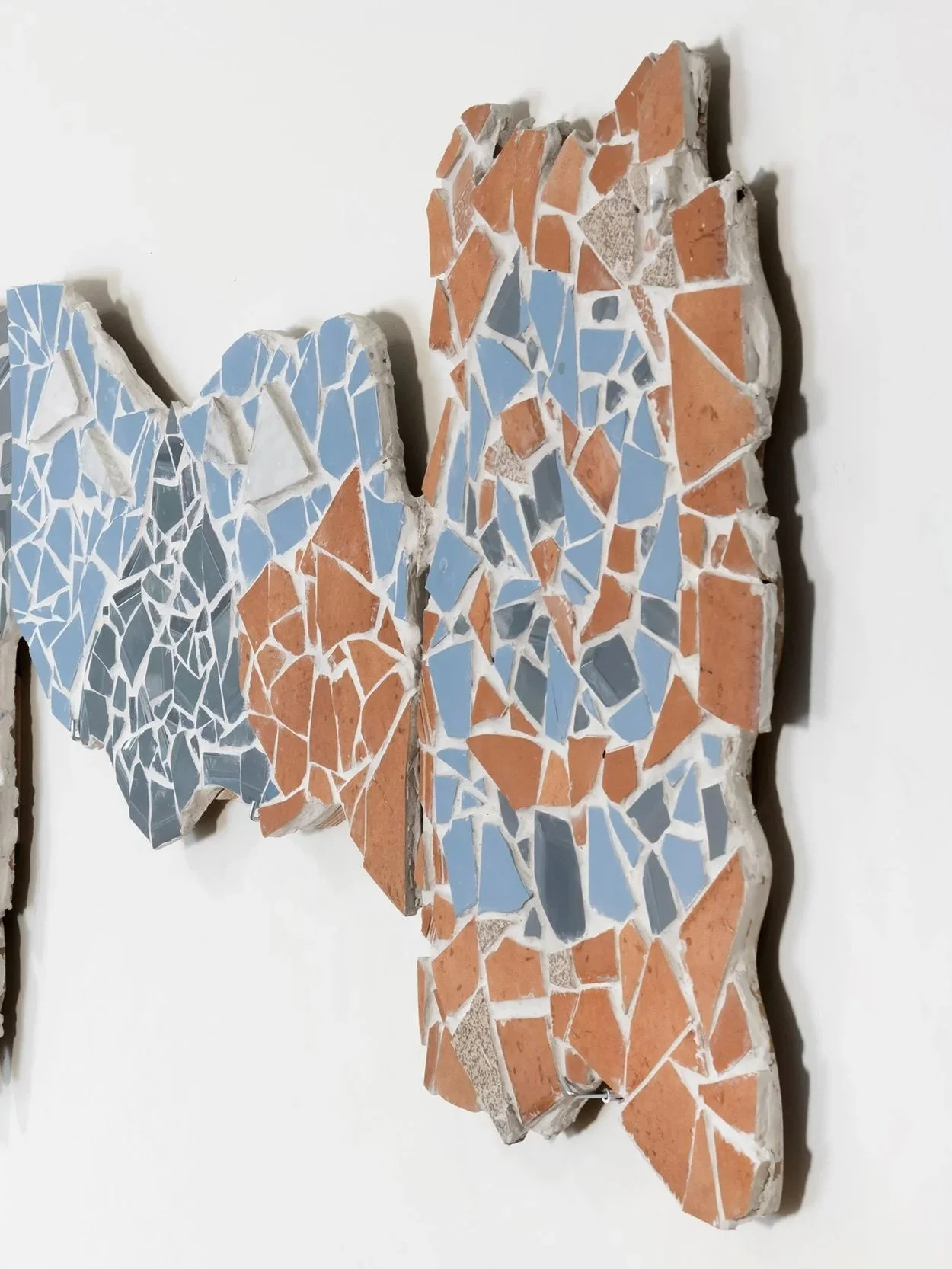 A mosaic art piece made of broken tiles in blue, orange, and gray colors on a white wall.