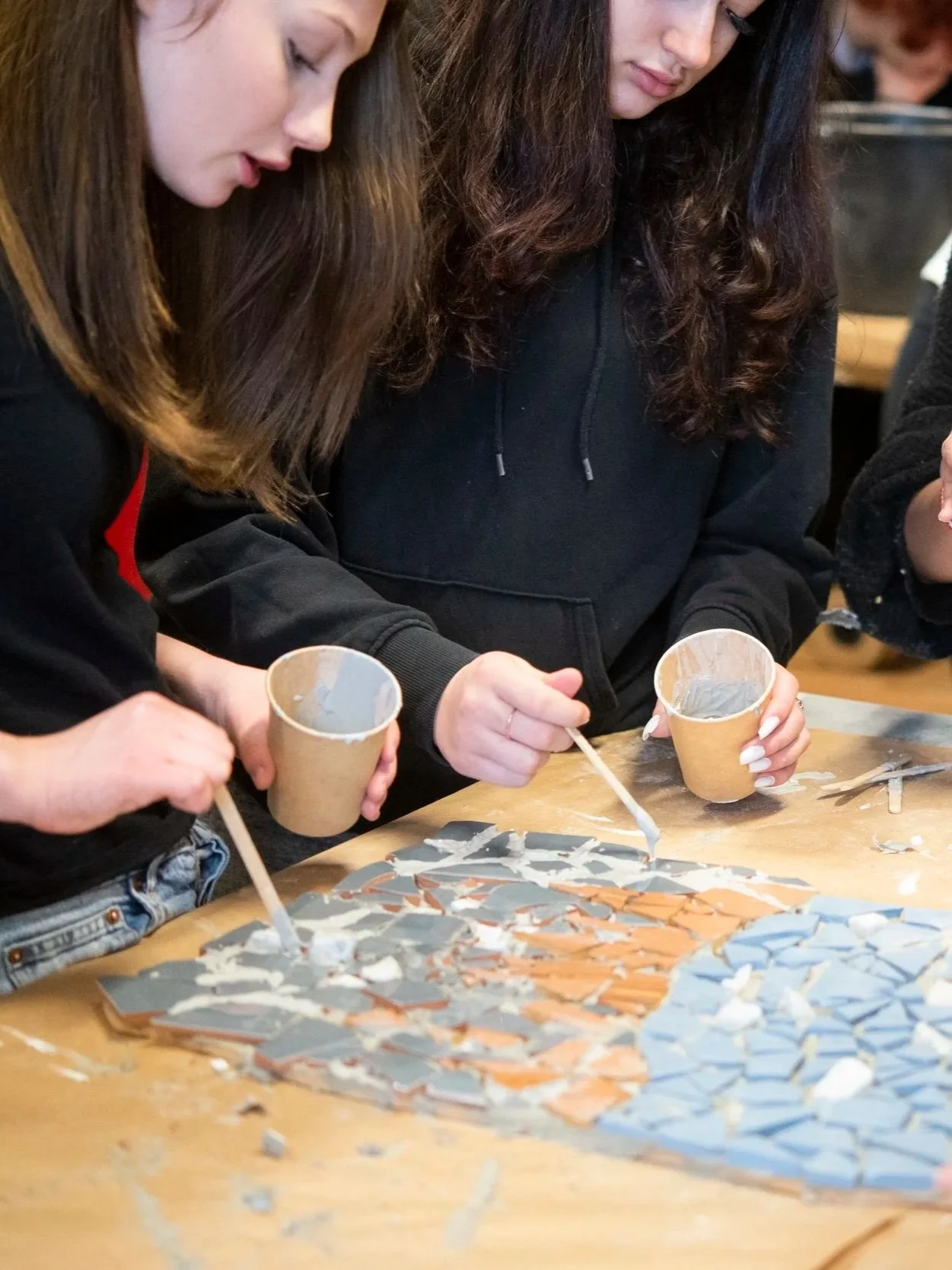 Two young women working on a mosaic project with broken tiles and cups of glue on a table.