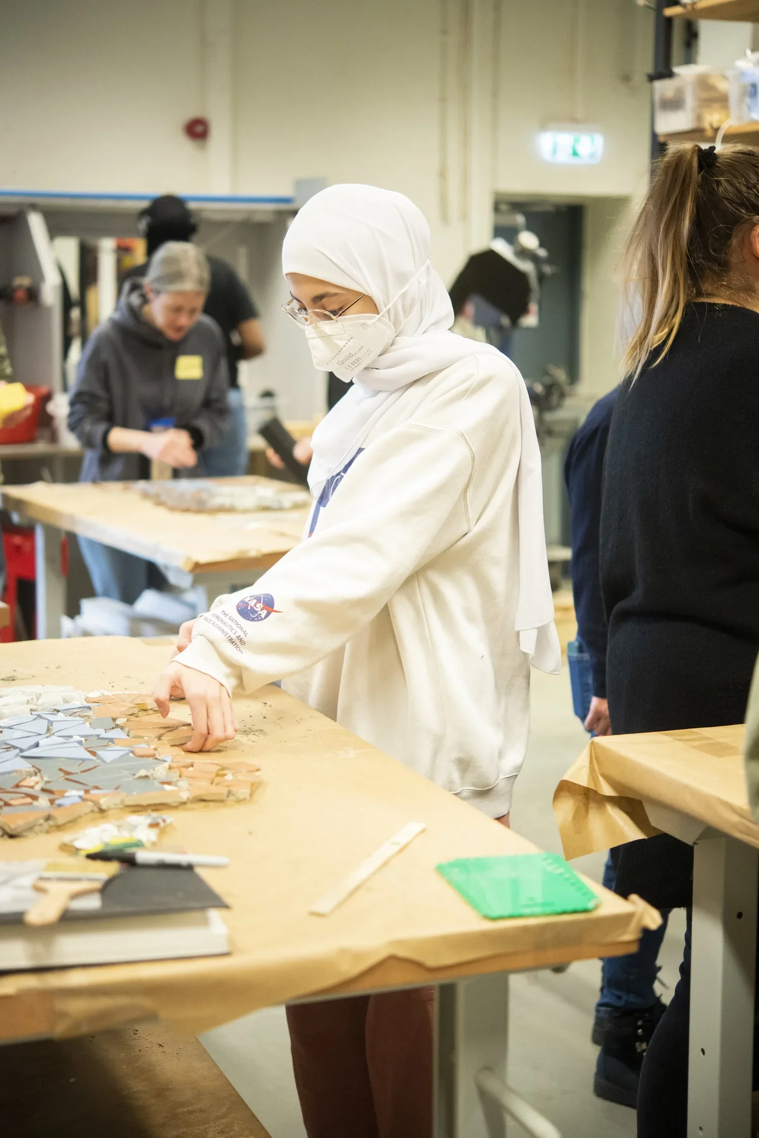 A woman wearing a white headscarf, face mask, and white hoodie working on a mosaic project at a workshop table, with other people working in the background.
