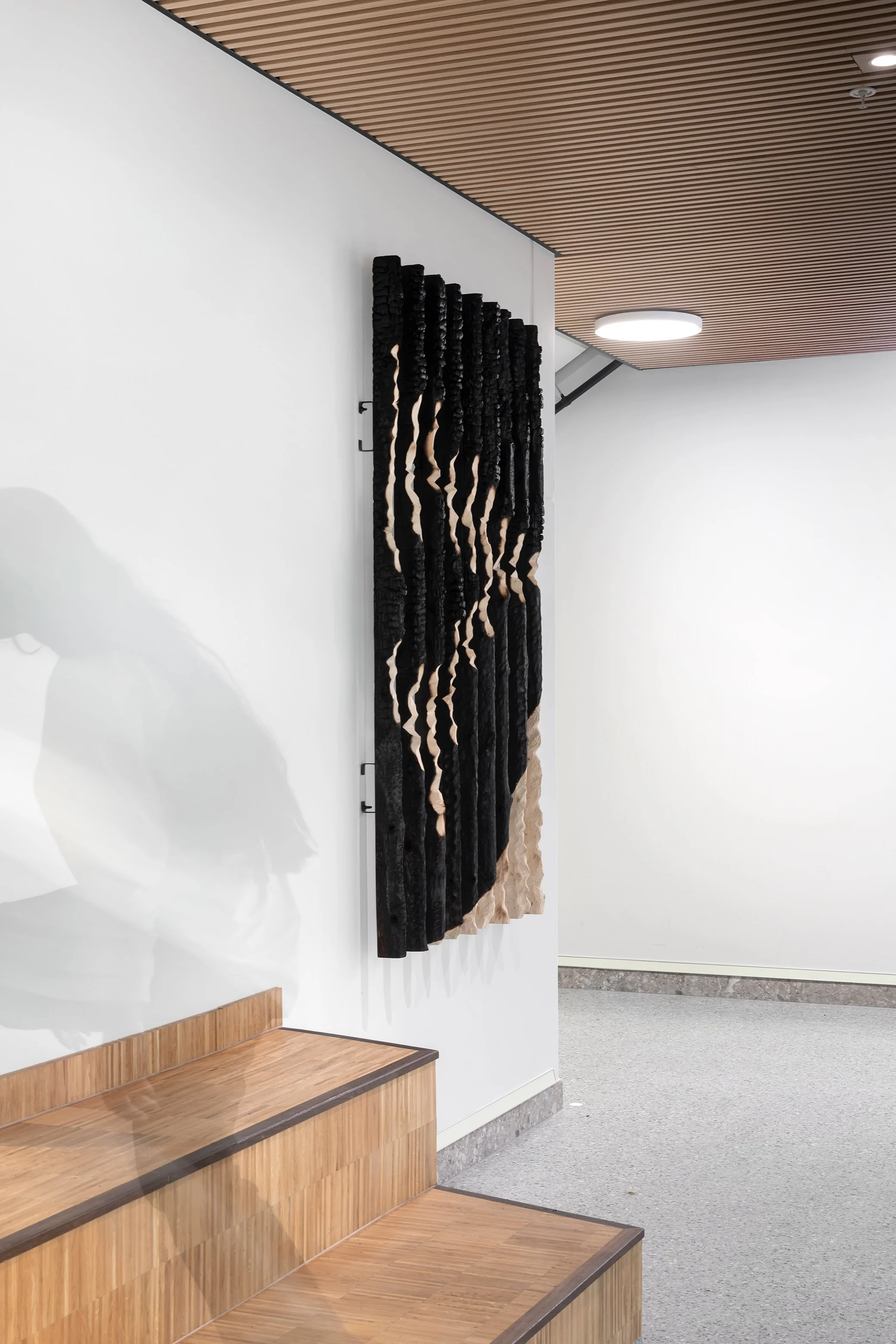 A modern interior wall display with dark and beige textured art pieces mounted vertically on a white wall. Wooden steps are positioned in the foreground, and a wooden ceiling with round ceiling lights is visible.