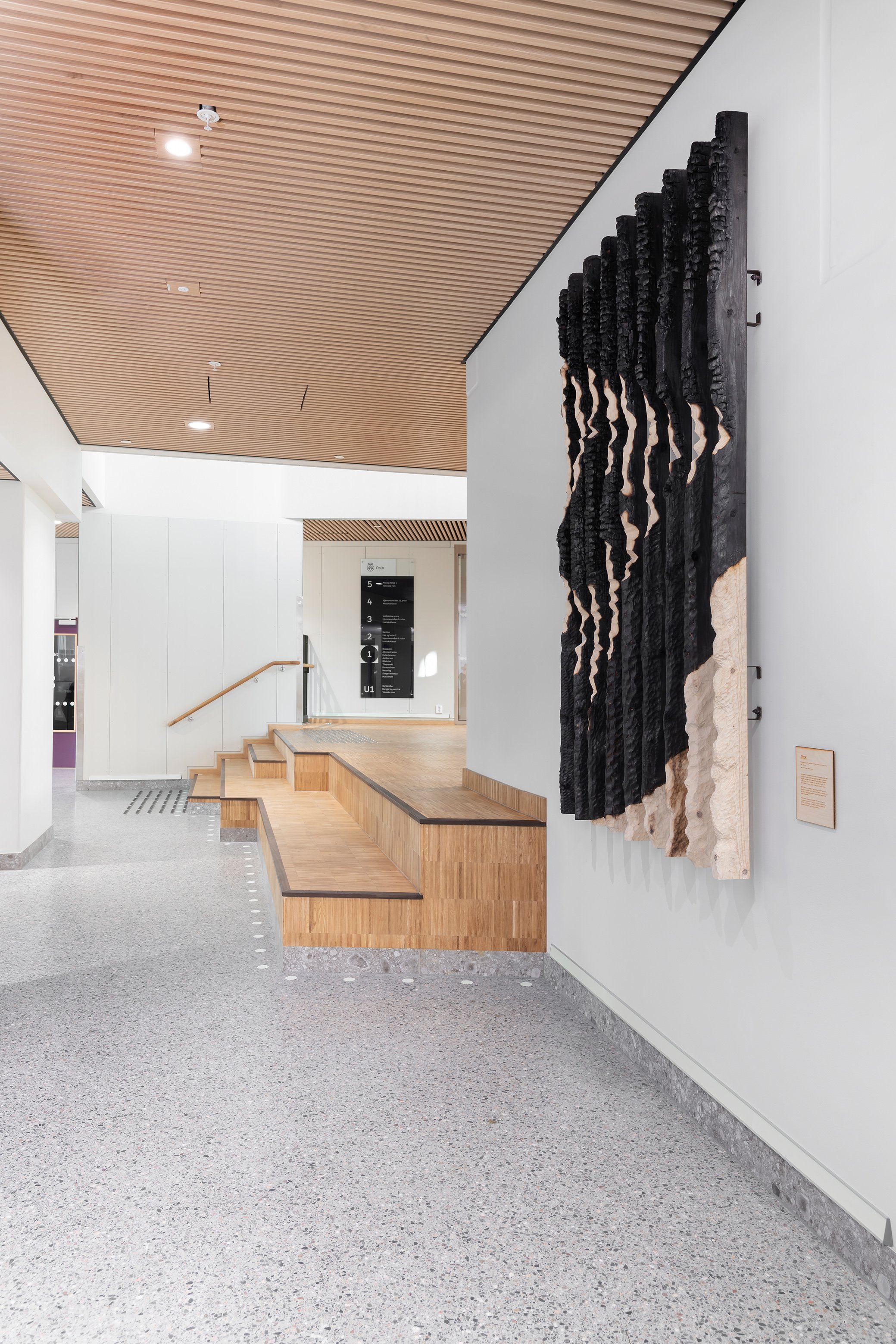 Modern interior with wooden ceiling, granite flooring, a wooden bench with steps, a textured wall art piece with black and beige twisted materials, and signage in the background.