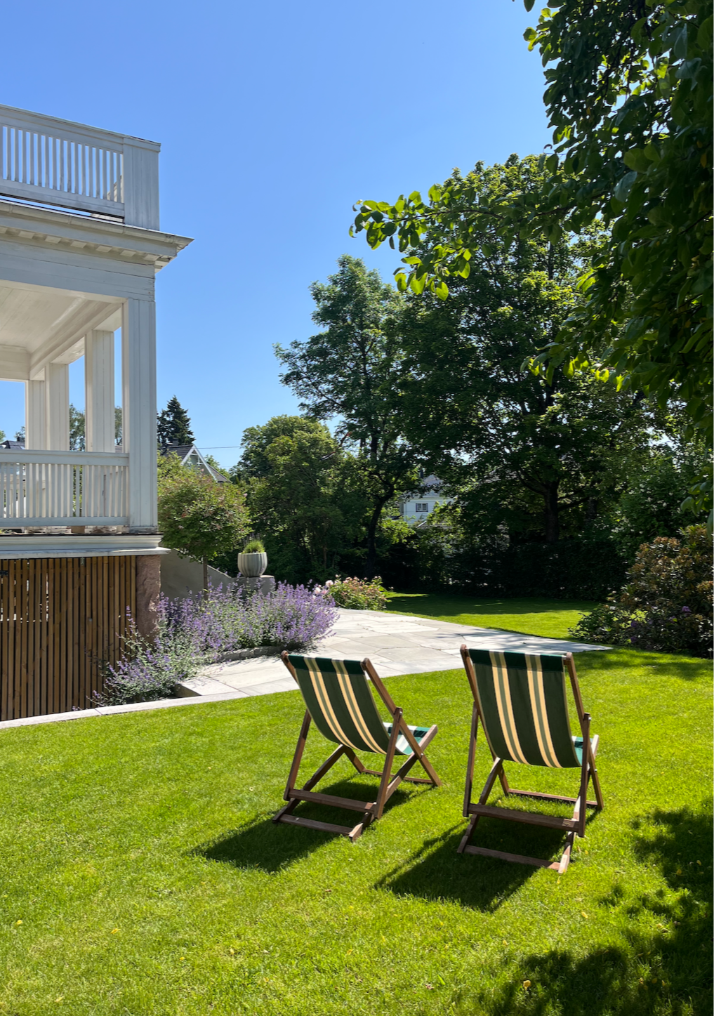 Two striped deck chairs on a well-manicured lawn in a backyard with lush trees, flowering bushes, and a bright blue sky.