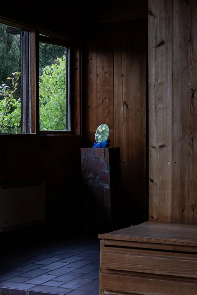 A corner of a wooden room with a window showing green trees outside, a small table with a mirror on top, and a wooden bench or chest.