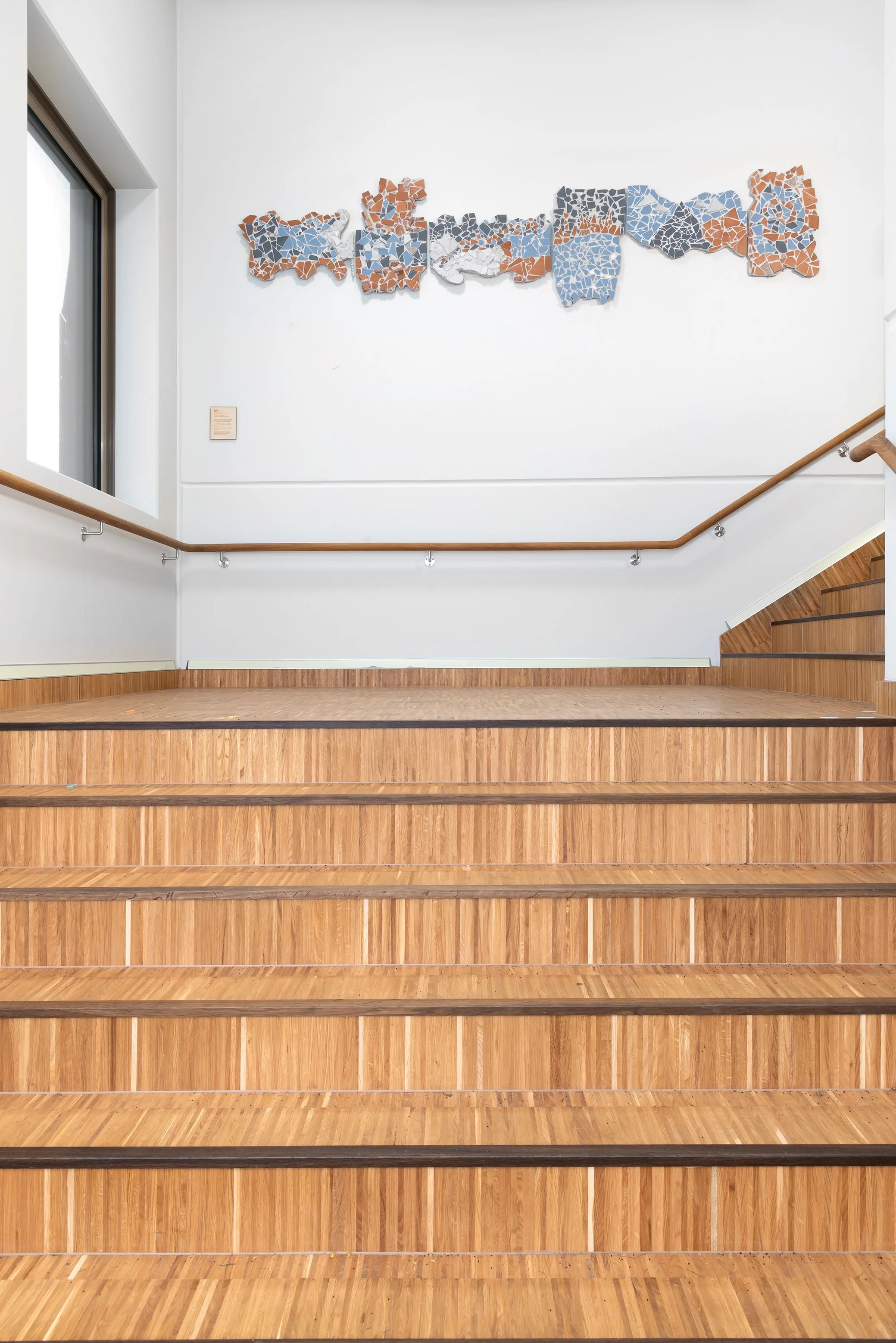 Wooden stairs leading up to a white wall with mosaic art and a window on the left.