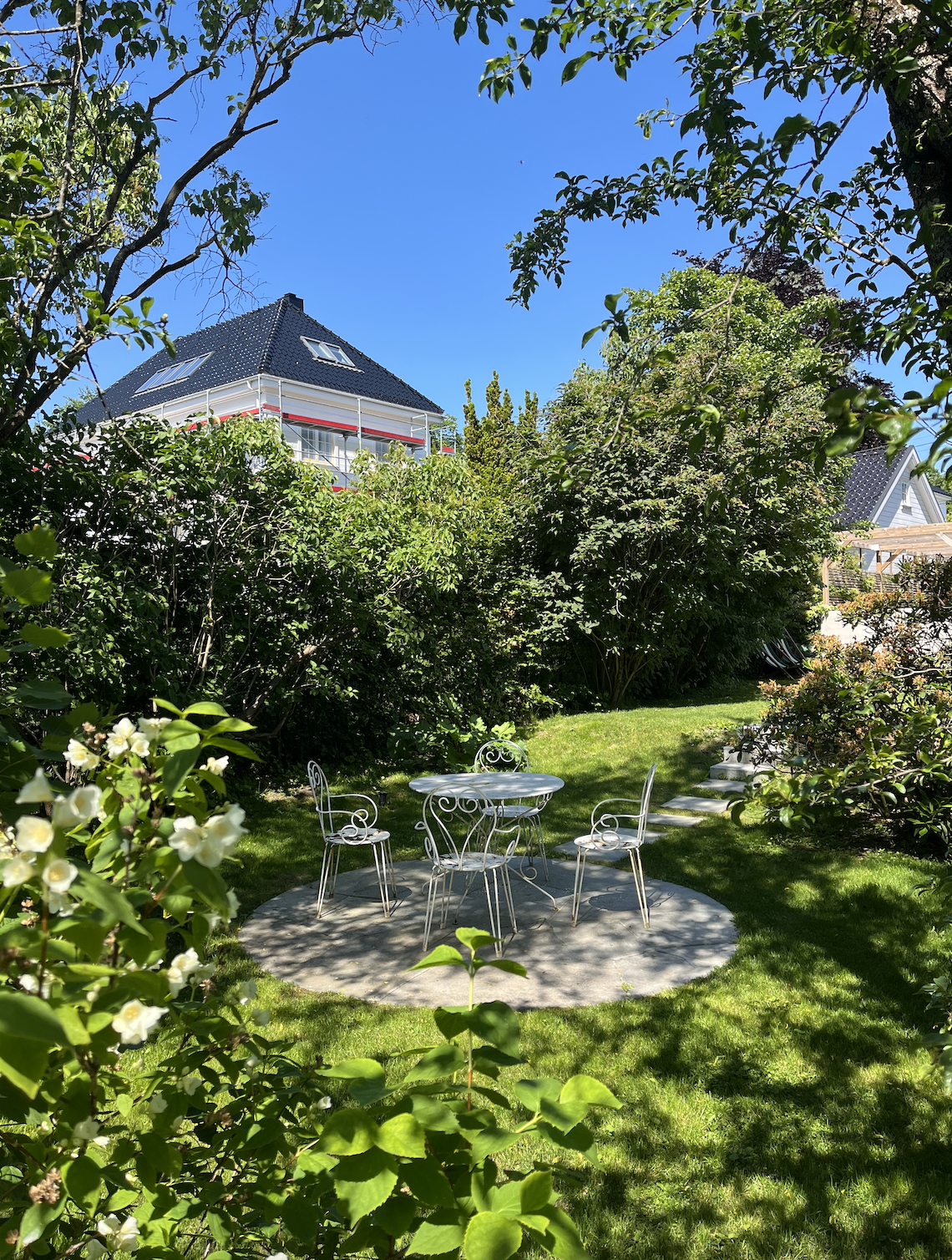 A backyard garden with a small white metal table and four matching chairs on a circular concrete pad, surrounded by lush green grass, trees, and bushes, with a house with a dark roof and a blue sky in the background.