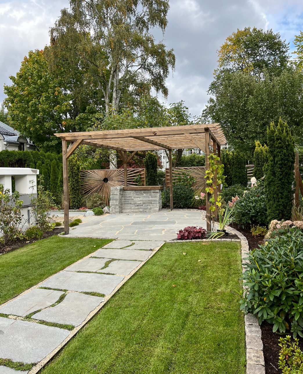 Backyard with a stone pathway, lush green lawn, and a wooden pergola surrounded by garden plants and trees under a partly cloudy sky.