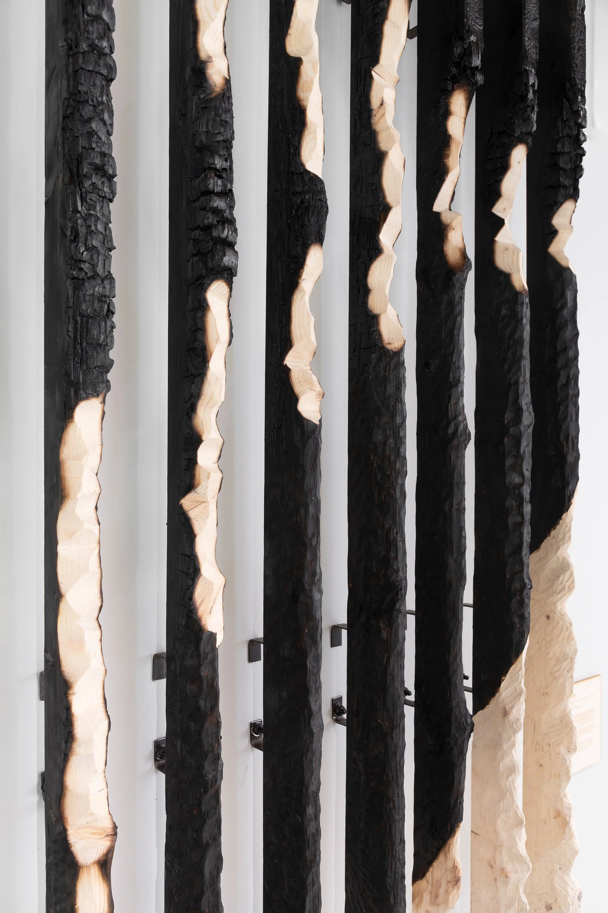 Vertical wooden art installation with charred and natural wood pieces, mounted on a white wall.