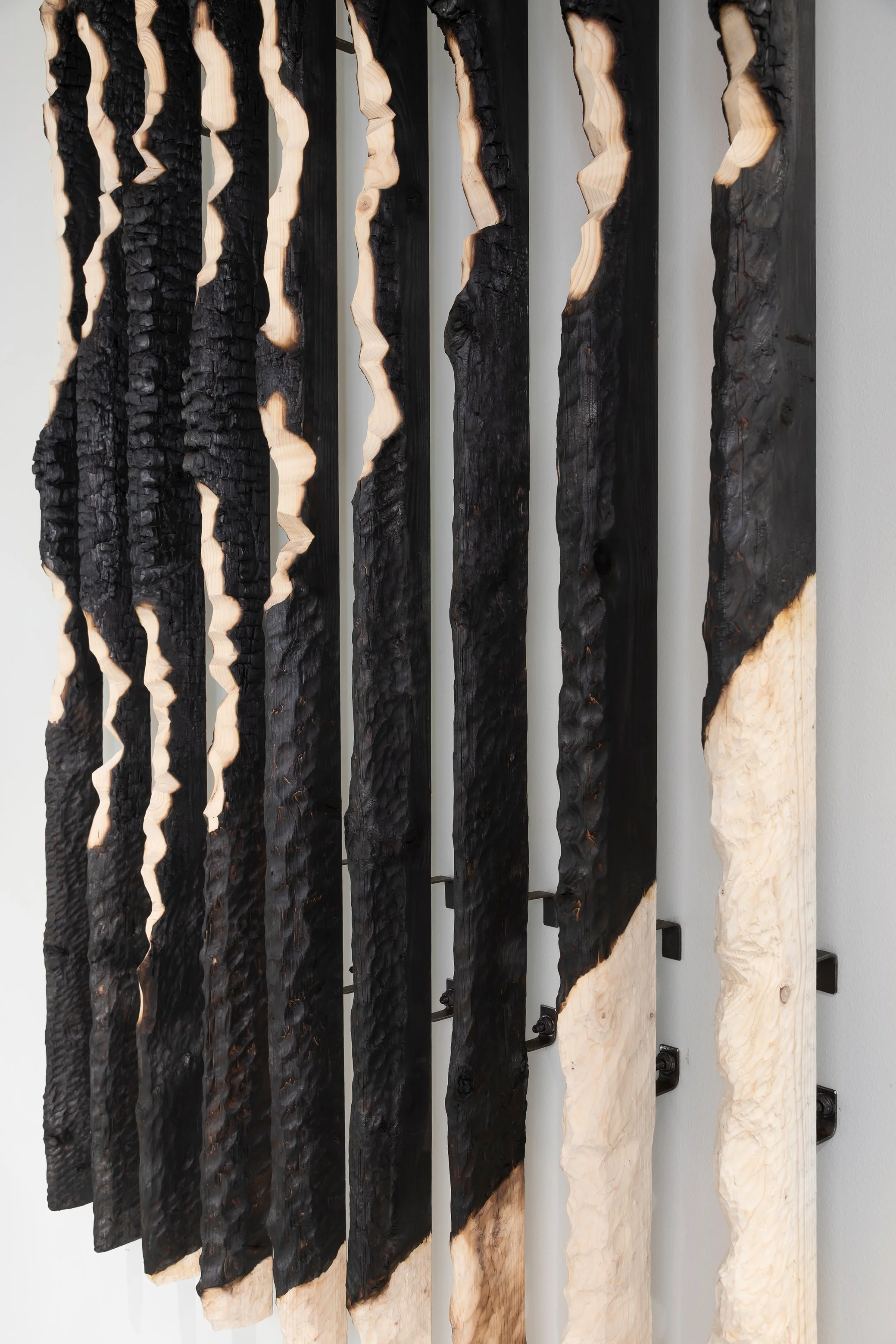 A close-up view of a modern wooden wall art installation made from vertically arranged wood planks with blackened sections, some with charred textures and others with natural light wood finish.