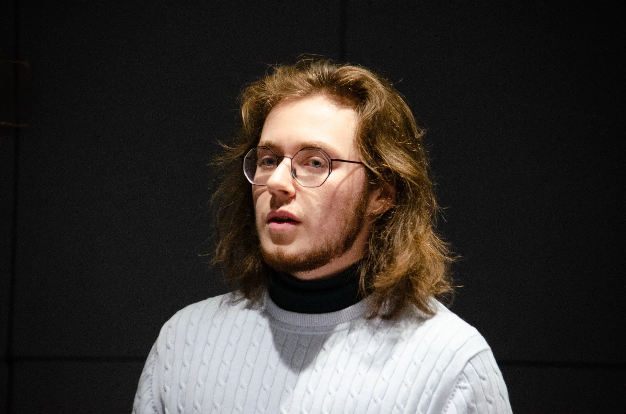 A photo of a white man with long wavy brown hair, wearing glasses. He looks pensive.