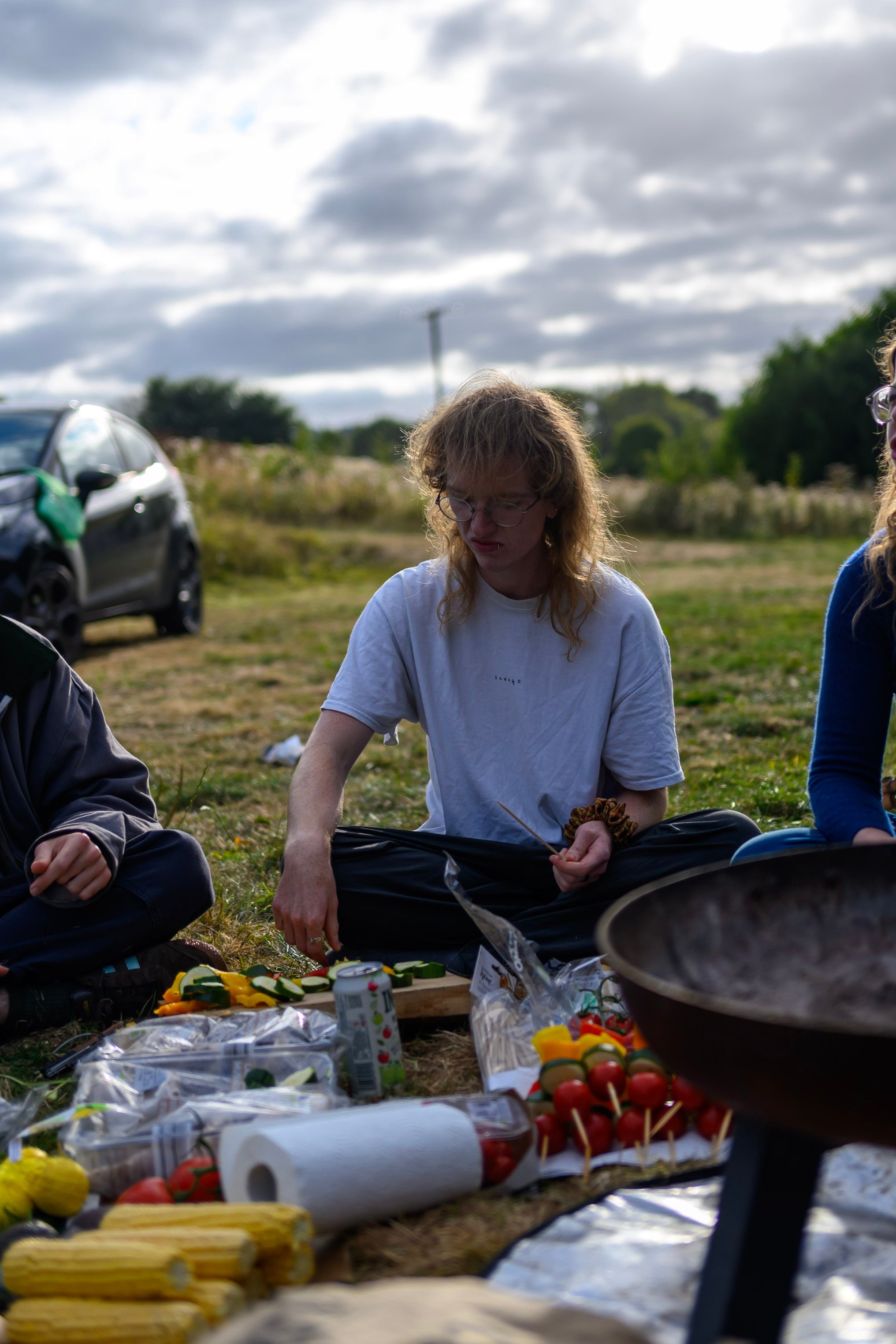 A photo of Kit, a skinny white person with a curly ginger mullet, sat on some grass outside. They are pulling a slightly comic face of disgust, looking at something on the ground