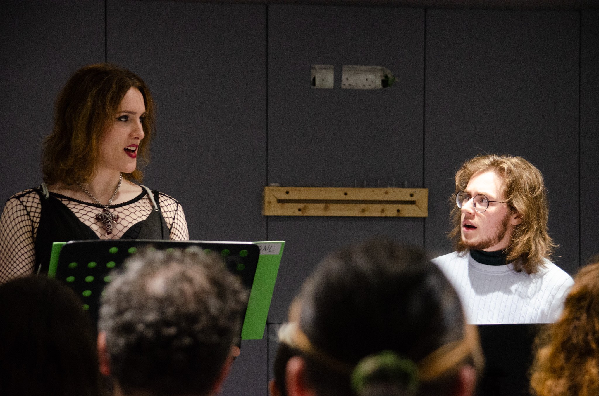 A photo of a tall woman and a short man standing in front of music stands, being watched by an audience. Both are white, with shoulder-length wavy brown hair, and they look a bit pissed off with each other.