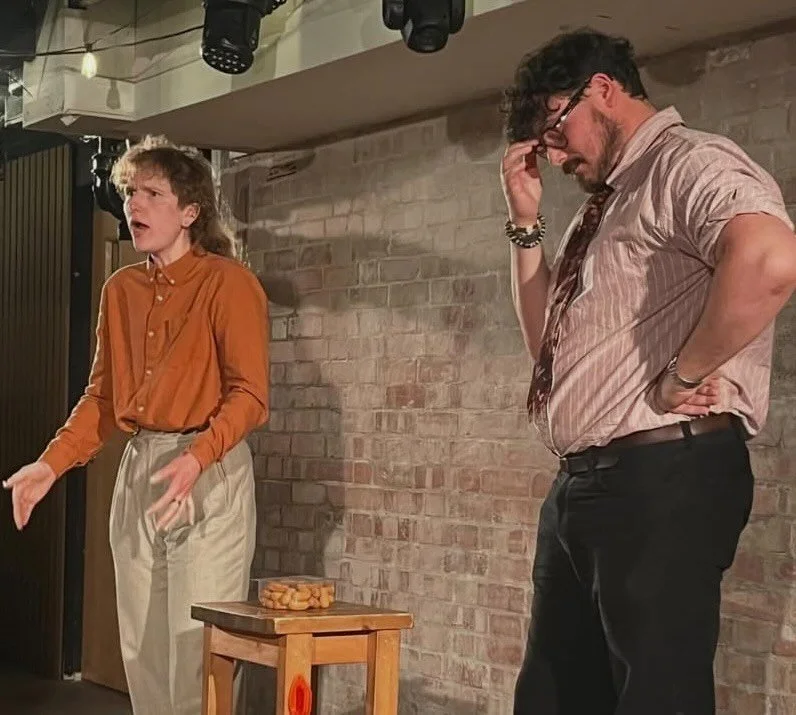 Kit, a skinny white person with a mullet, and Alex, a larger dark-haired man, stand on stage wearing business attire. A box of cocktail sausages sits between them. Kit is ranting, Alex has his head in his hands.
