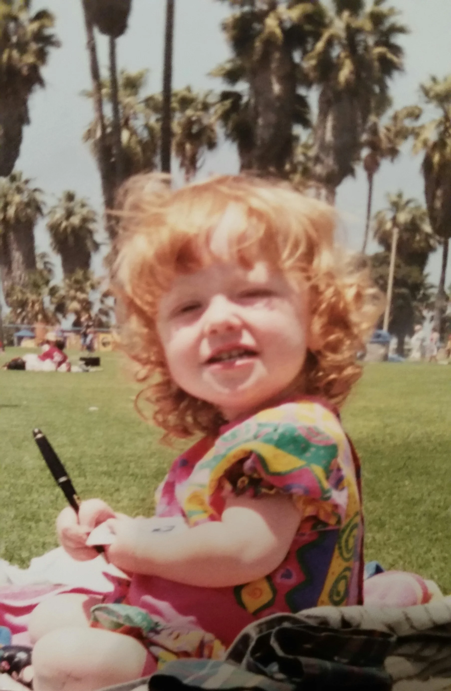 A photo of Kit as a toddler. They have curly red hair to their shoulders and a colourful top. They are looking at the camera, holding a pen and paper, gripping the pen like an adult.