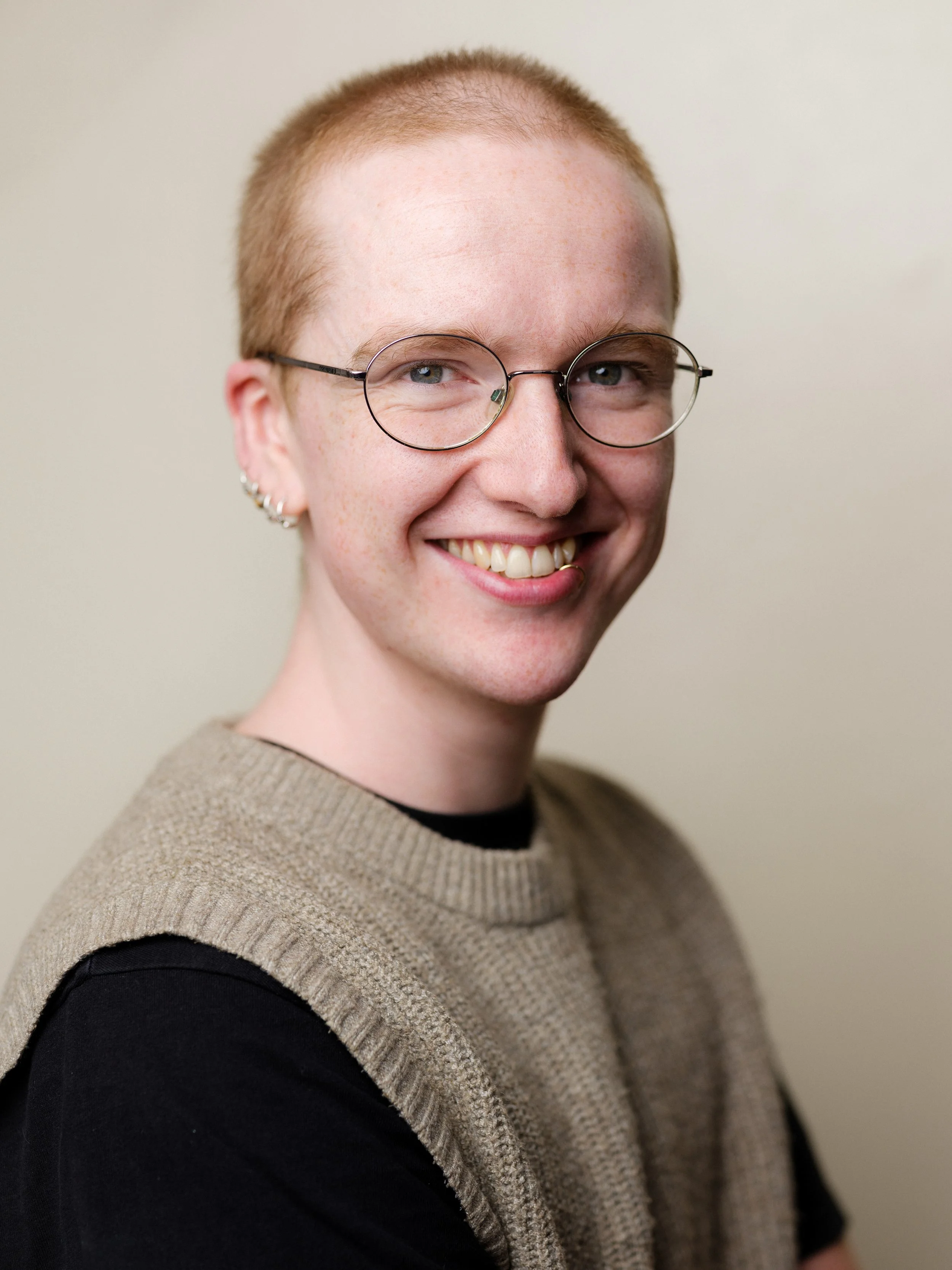 A headshot of Kit McGuire: a white nonbinary peson with a ginger buzzcut. They are smiling at the camera and wearing round glasses.