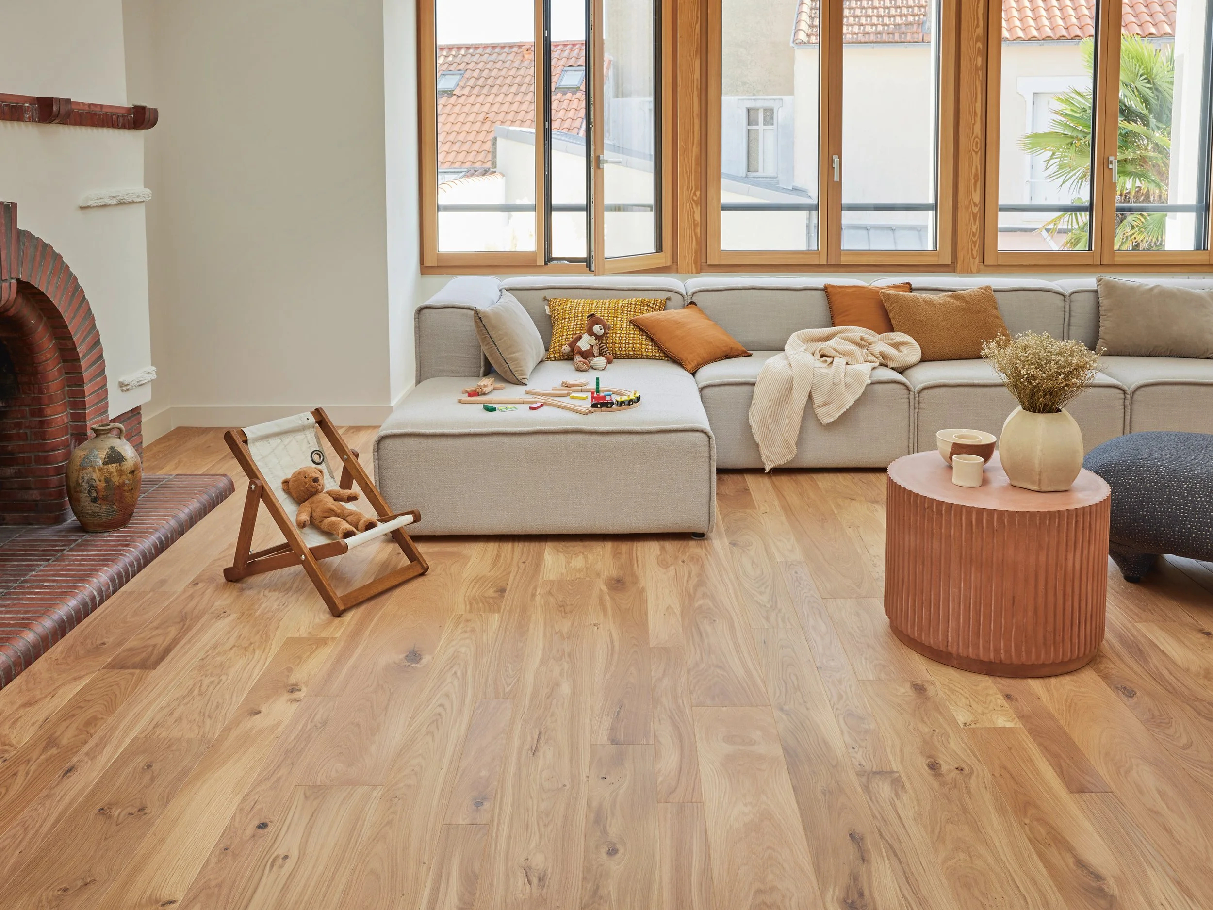 Bright living room with wooden floors, a large off-white sectional sofa with brown and grey cushions, and toys on it. A small wooden folding chair with a teddy bear, a brick fireplace on the left, and a round pink textured coffee table with a vase of dried flowers and some bowls. Large windows with wooden frames let in natural light, showing rooftops and a palm tree outside.