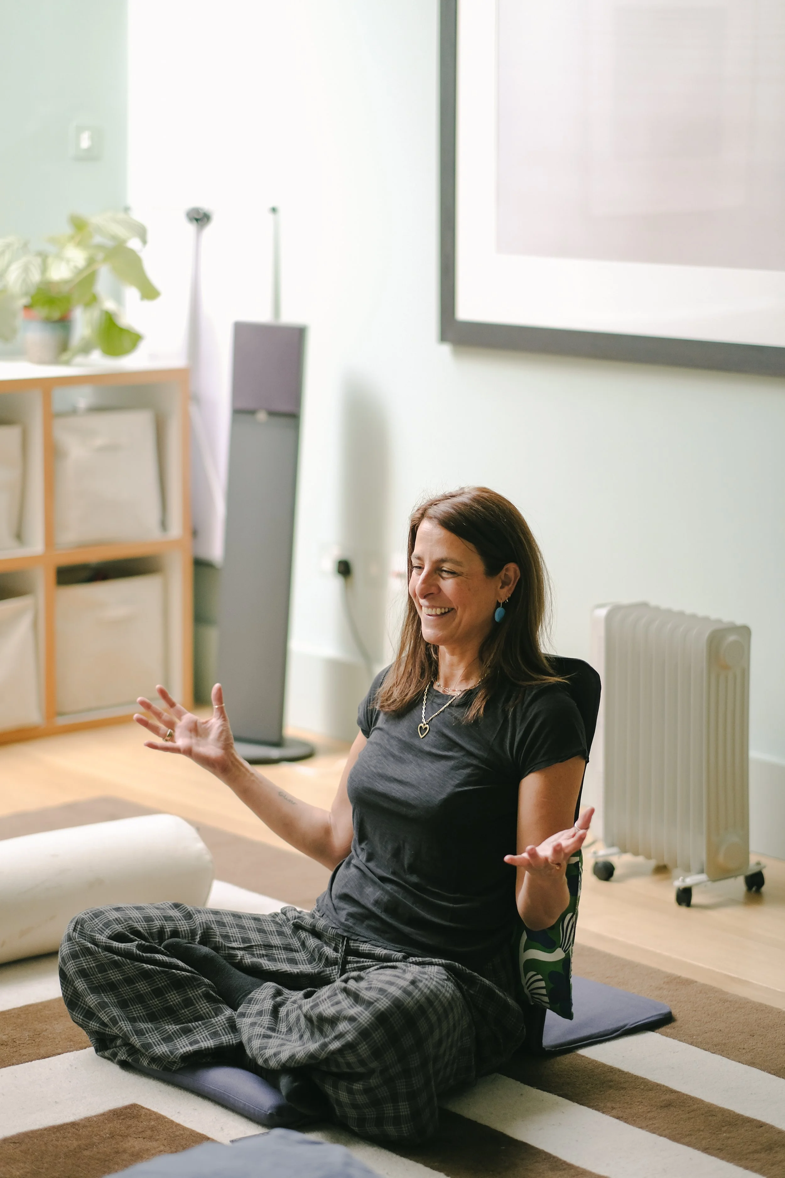 Woman sitting on a yoga mat with legs crossed, smiling and gesturing with hands in a room with a striped rug, a filing cabinet with storage bins, potted plant, and a radiator.