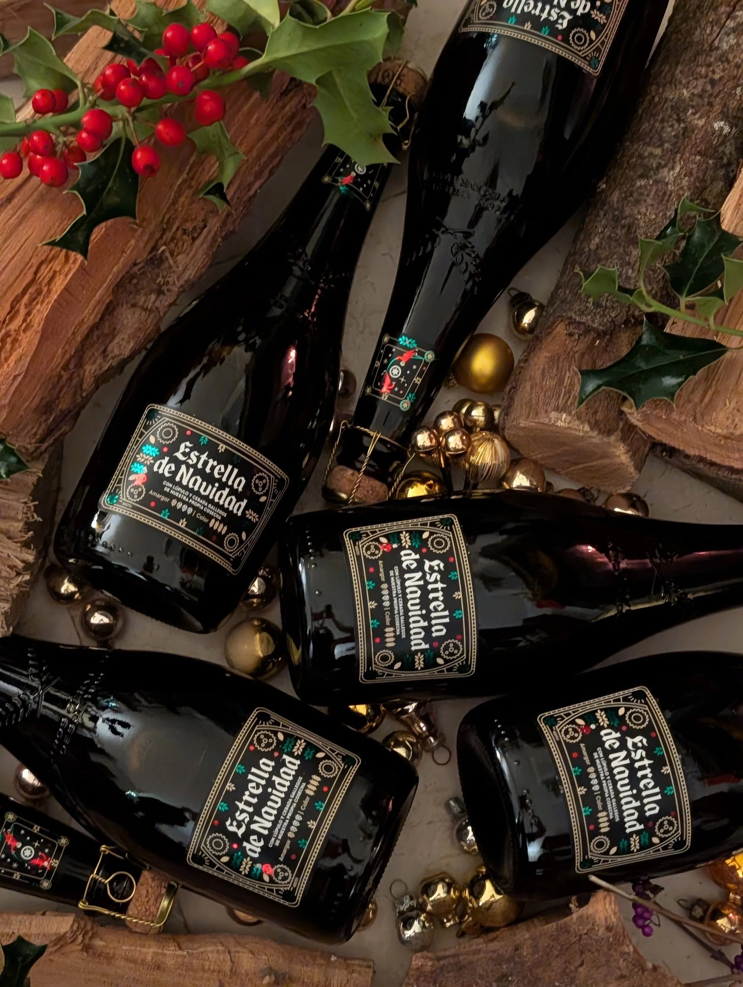 Four dark glass bottles labeled 'Estrella de Navidad' surrounded by gold and metallic ornaments, greenery, and wooden logs.