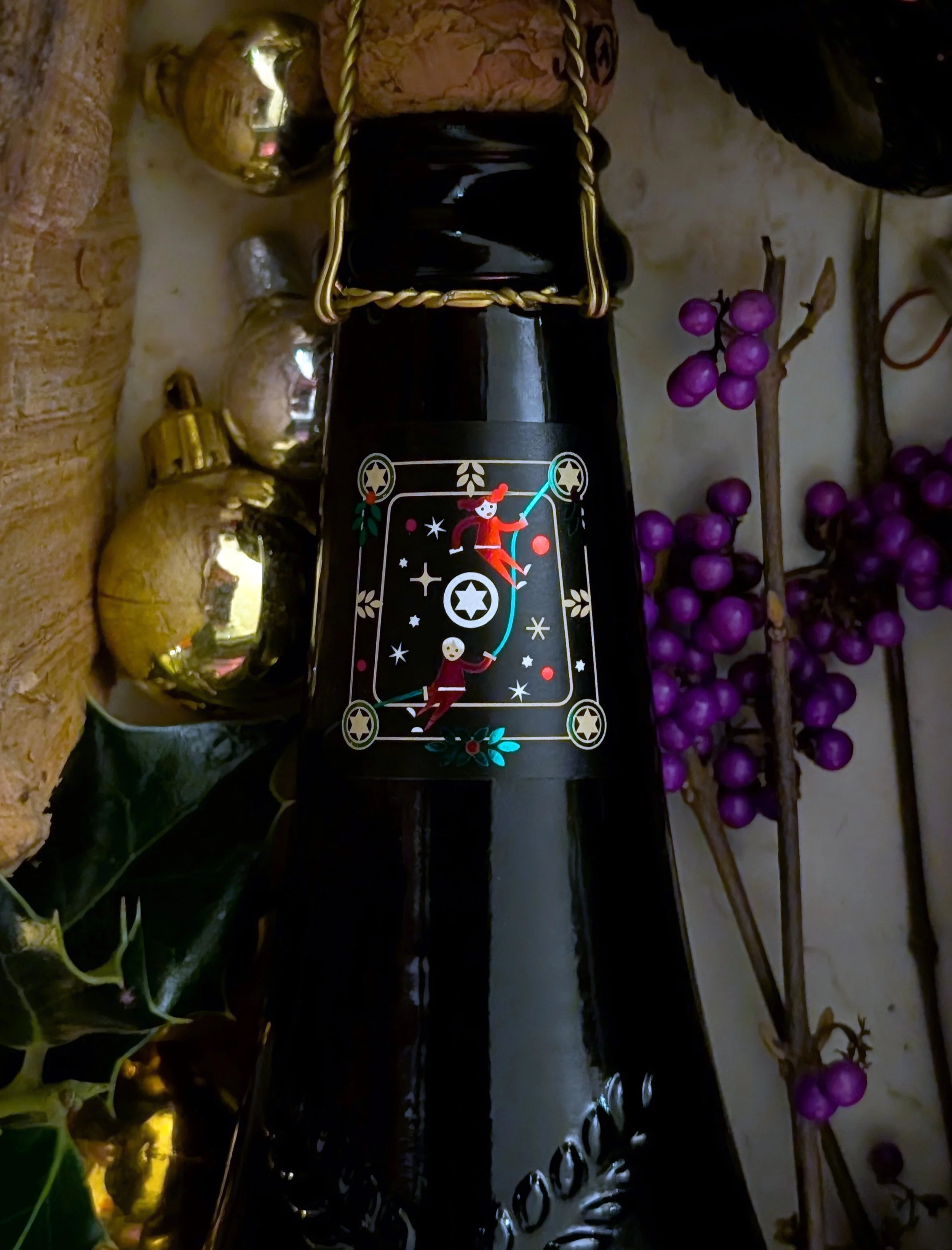 A black bottle decorated with a mystical tarot card design, surrounded by gold and silver ornaments, and purple berries on branches, on a wooden surface.