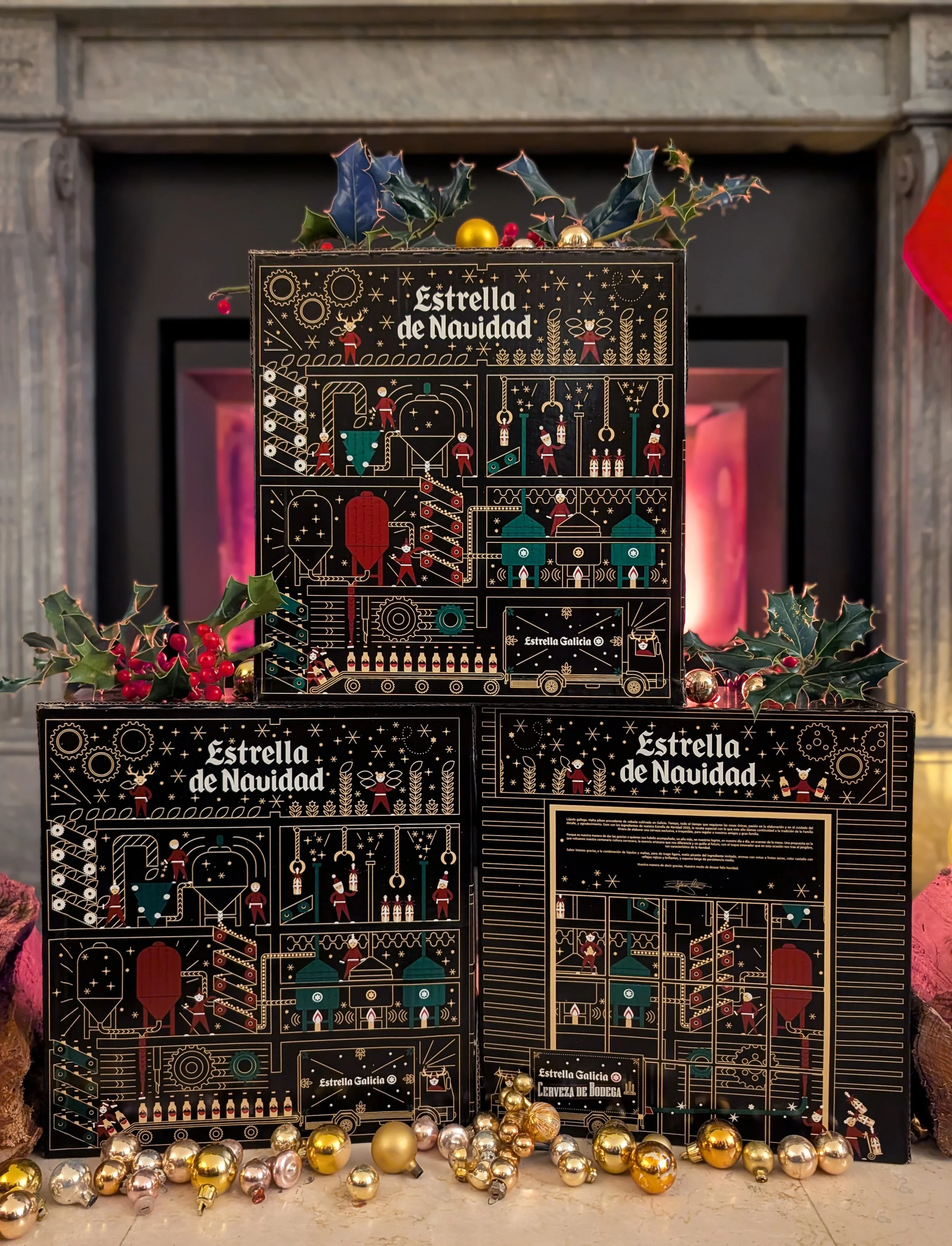 Three black boxes with gold Christmas-themed illustrations and the words "Estrella de Navidad" stacked on a fireplace mantle, decorated with Christmas ornaments, holly leaves, and berries.